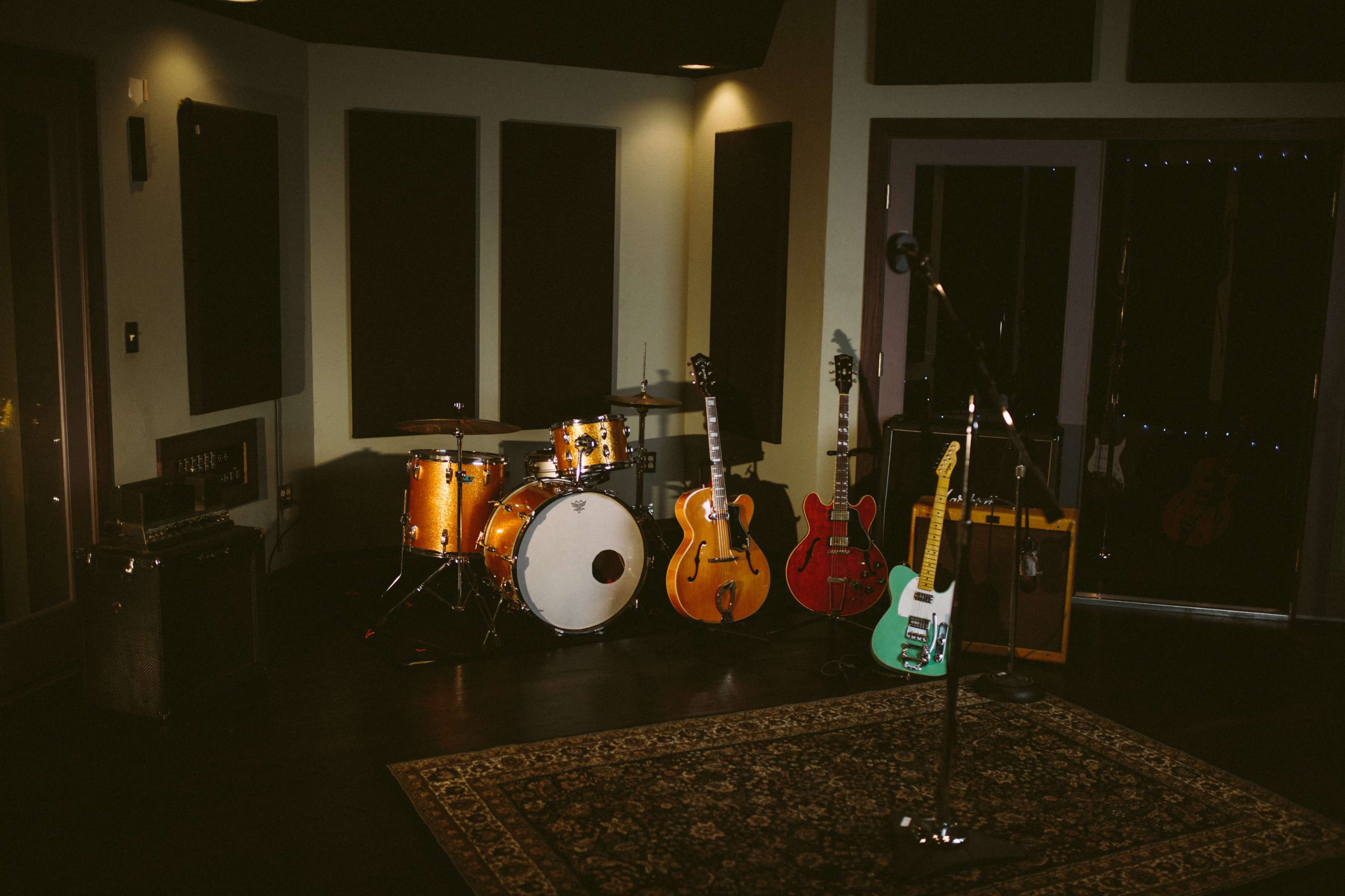 A music studio with a drum set, several guitars, and amplifiers arranged around a microphone stand on a patterned rug.