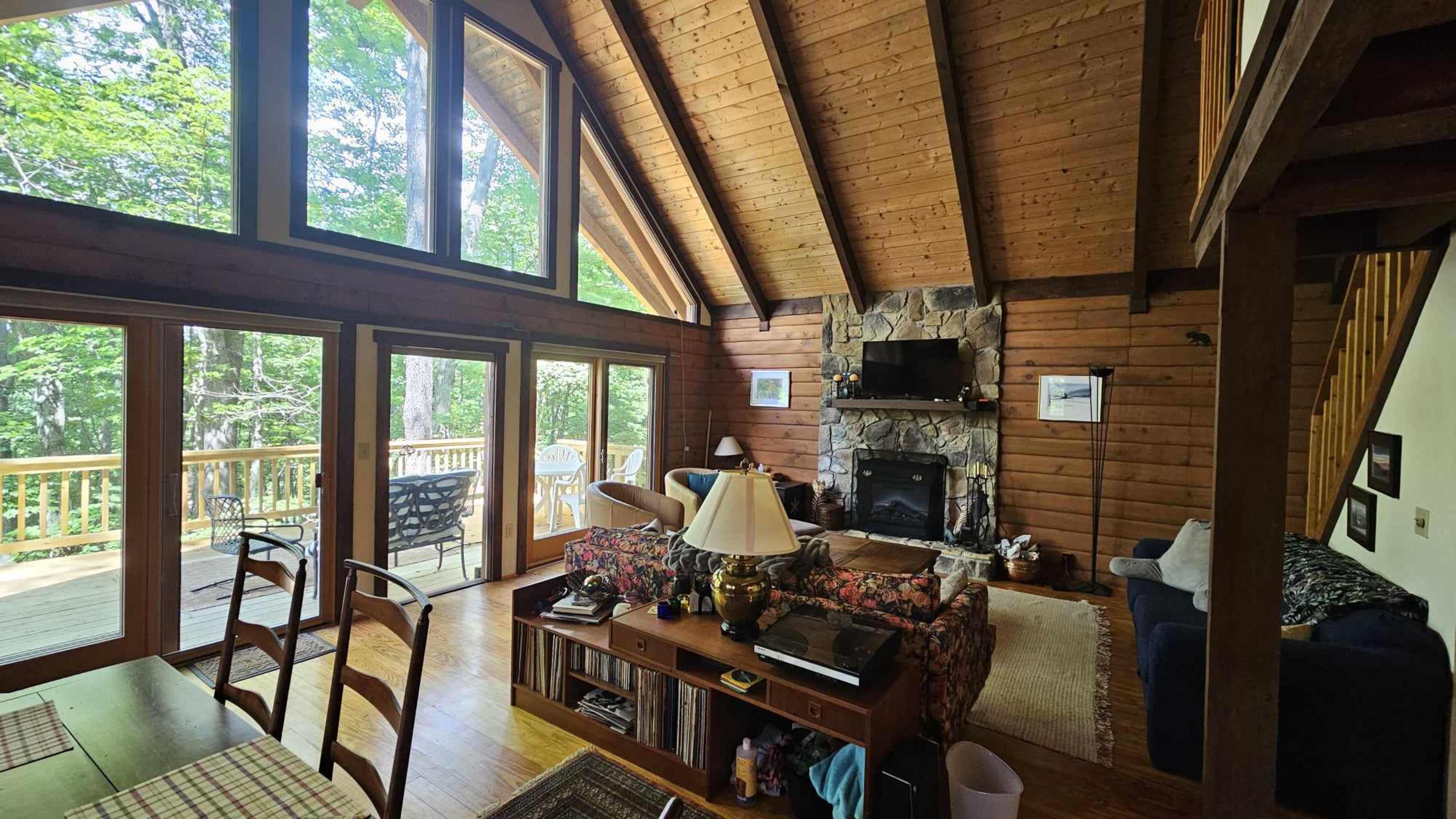 The interior of a wooden cabin features large windows, a stone fireplace, and a cozy seating area with a mix of tables and decor.