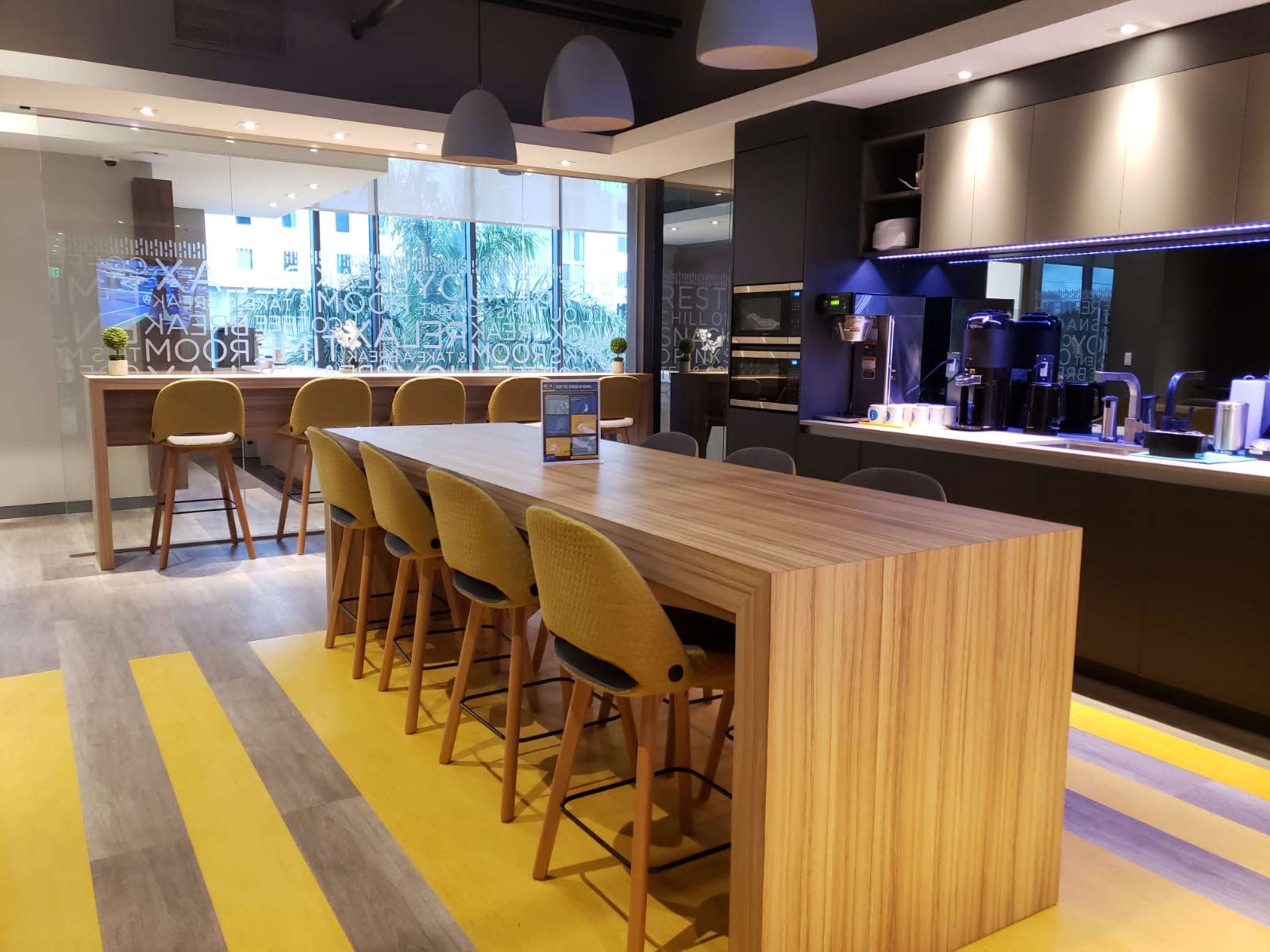 A modern kitchen space featuring a long wooden table surrounded by high-backed stools, with appliances and a bar area in the background.