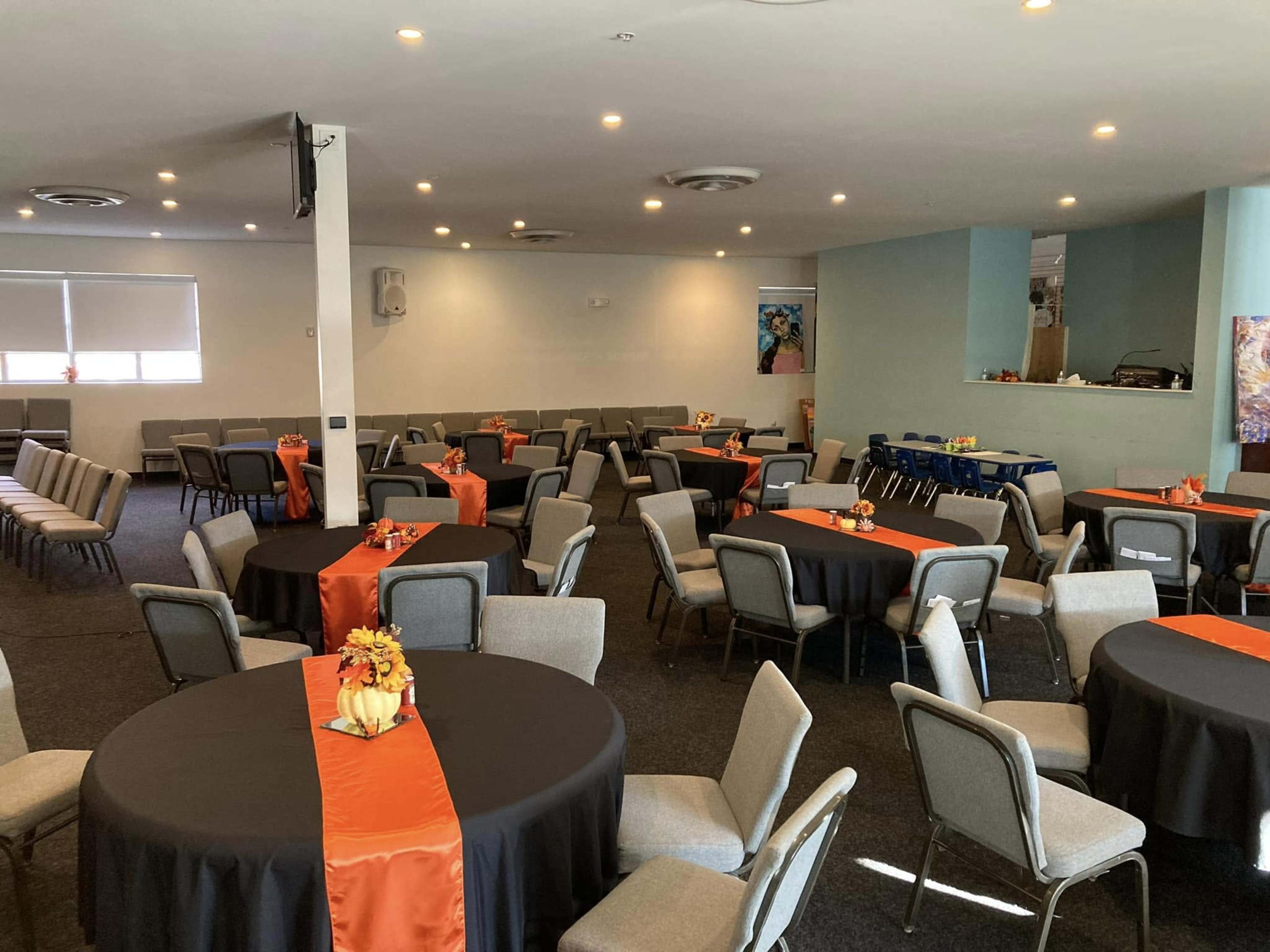 A large room set up for an event featuring round tables covered with black tablecloths and orange runners, surrounded by gray chairs.
