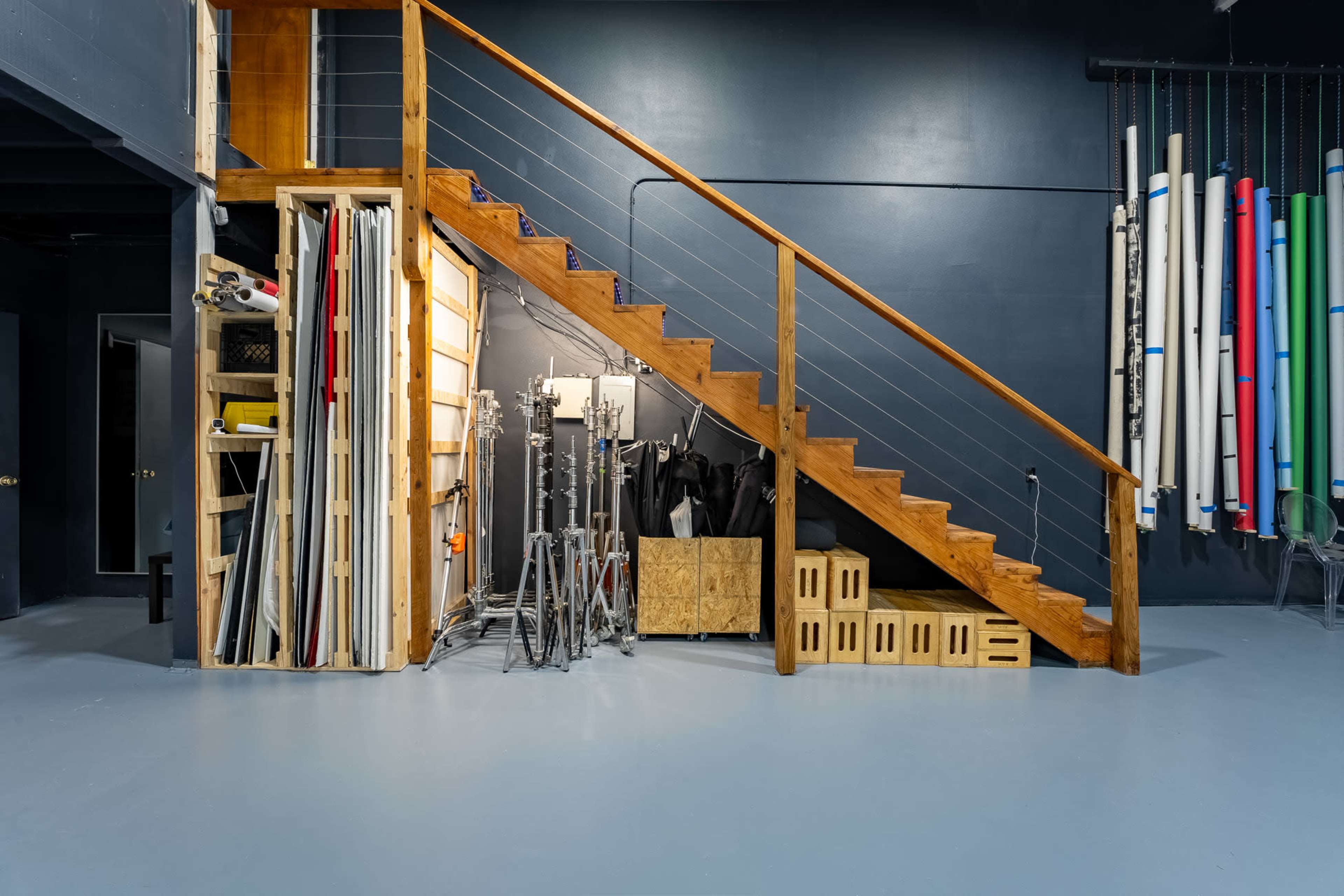 A studio space with wooden stairs leading to a higher level, alongside organized photography equipment and rolls of backdrop materials.