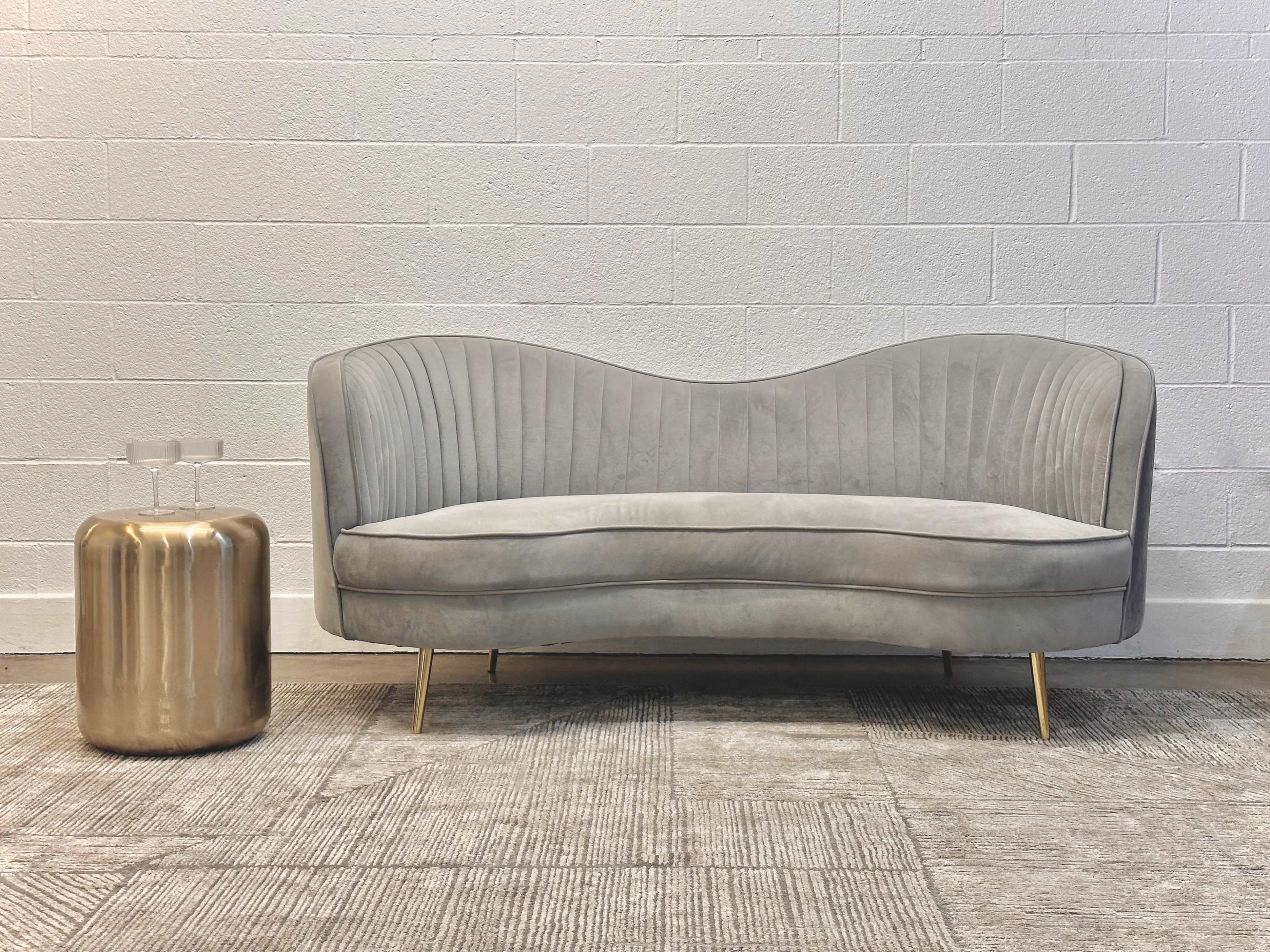 A gray velvet sofa with curved edges is positioned next to a golden, cylindrical side table on a textured floor.