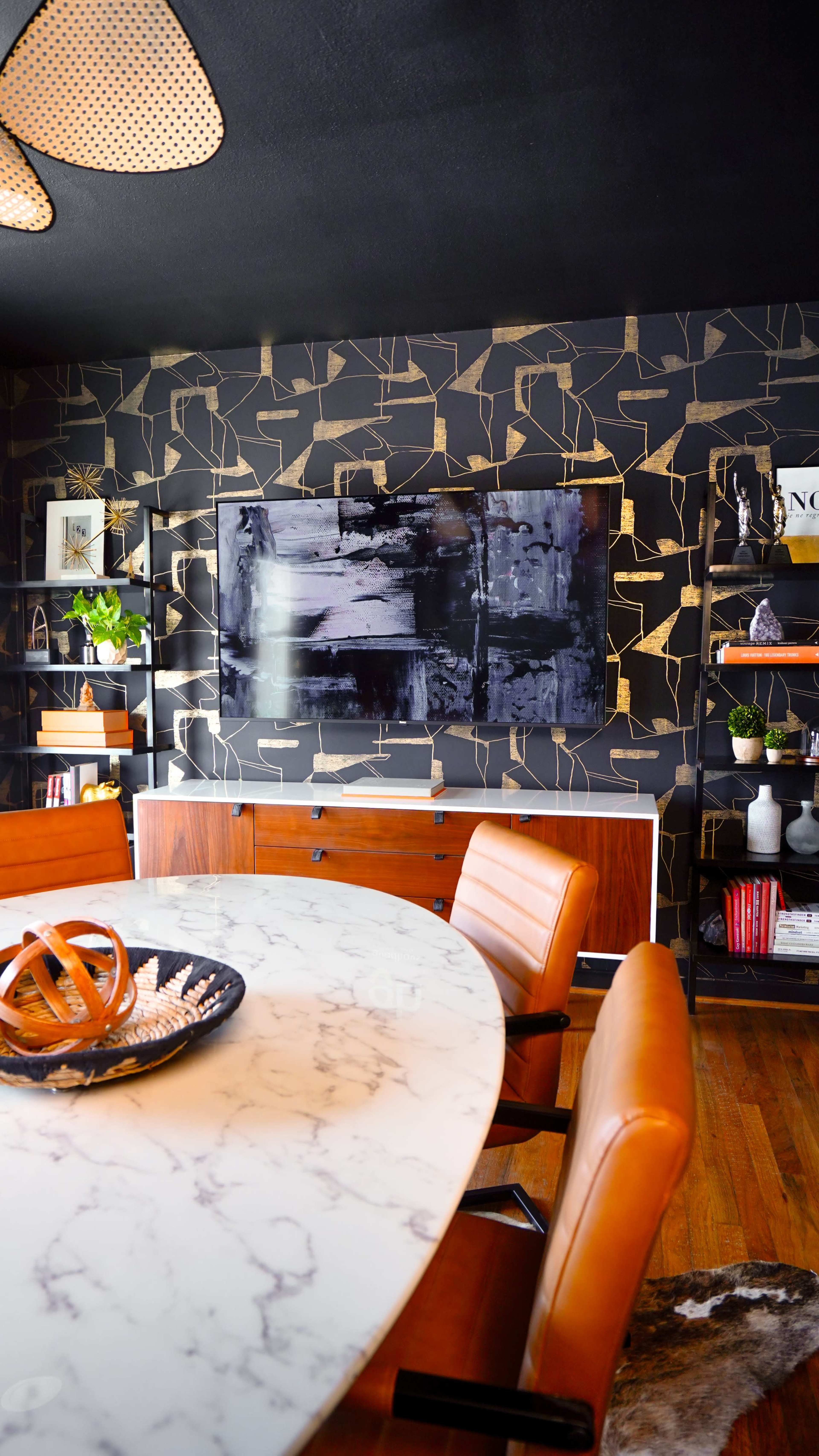 The image shows a modern dining area featuring a round marble table with wooden chairs, a television mounted on a gold-accented black wall, and open shelving displaying decorative items and plants.