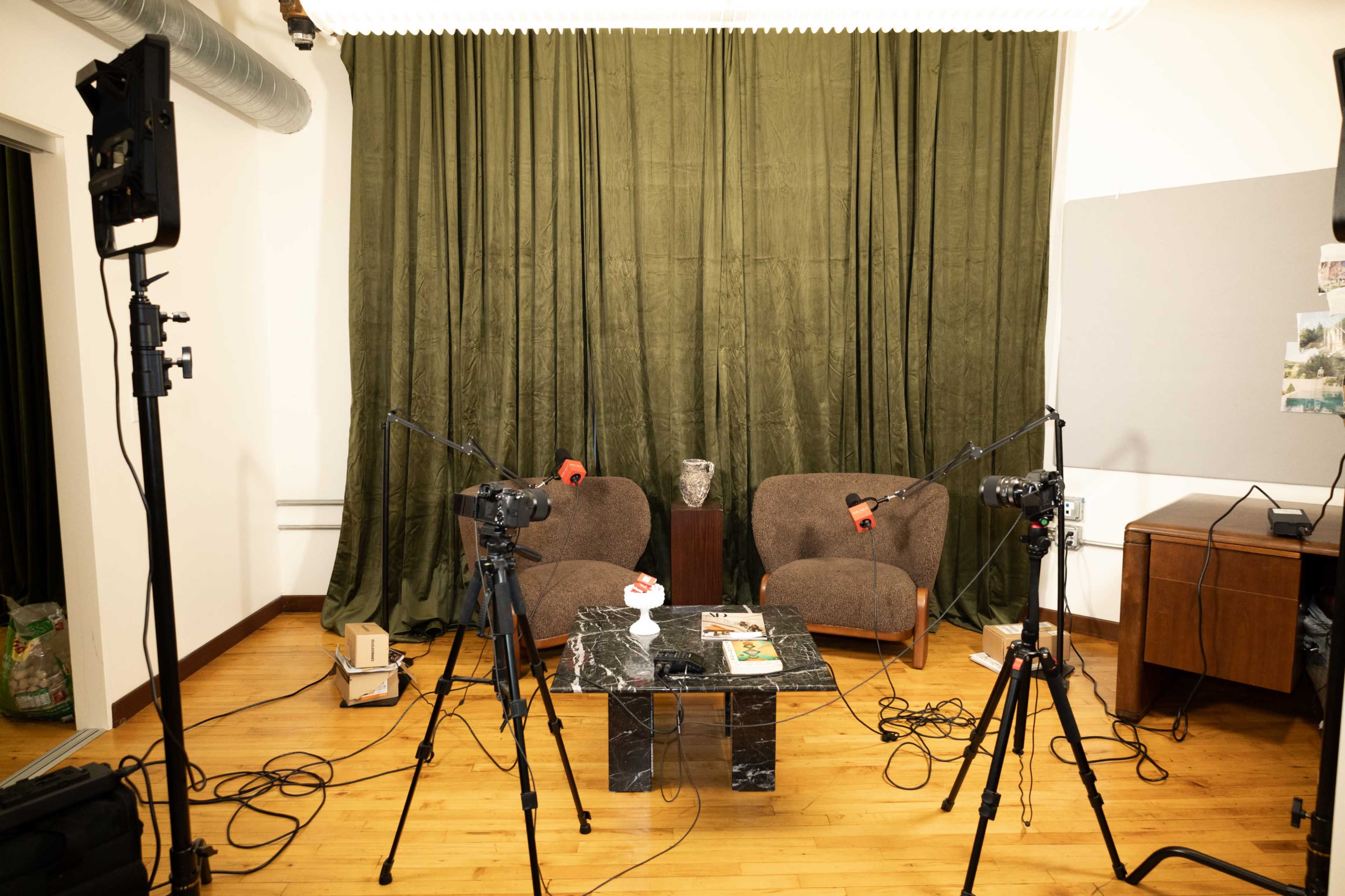 The image shows a studio setup with two brown chairs facing a small black marble table, surrounded by camera tripods and microphones, against a green curtain backdrop.