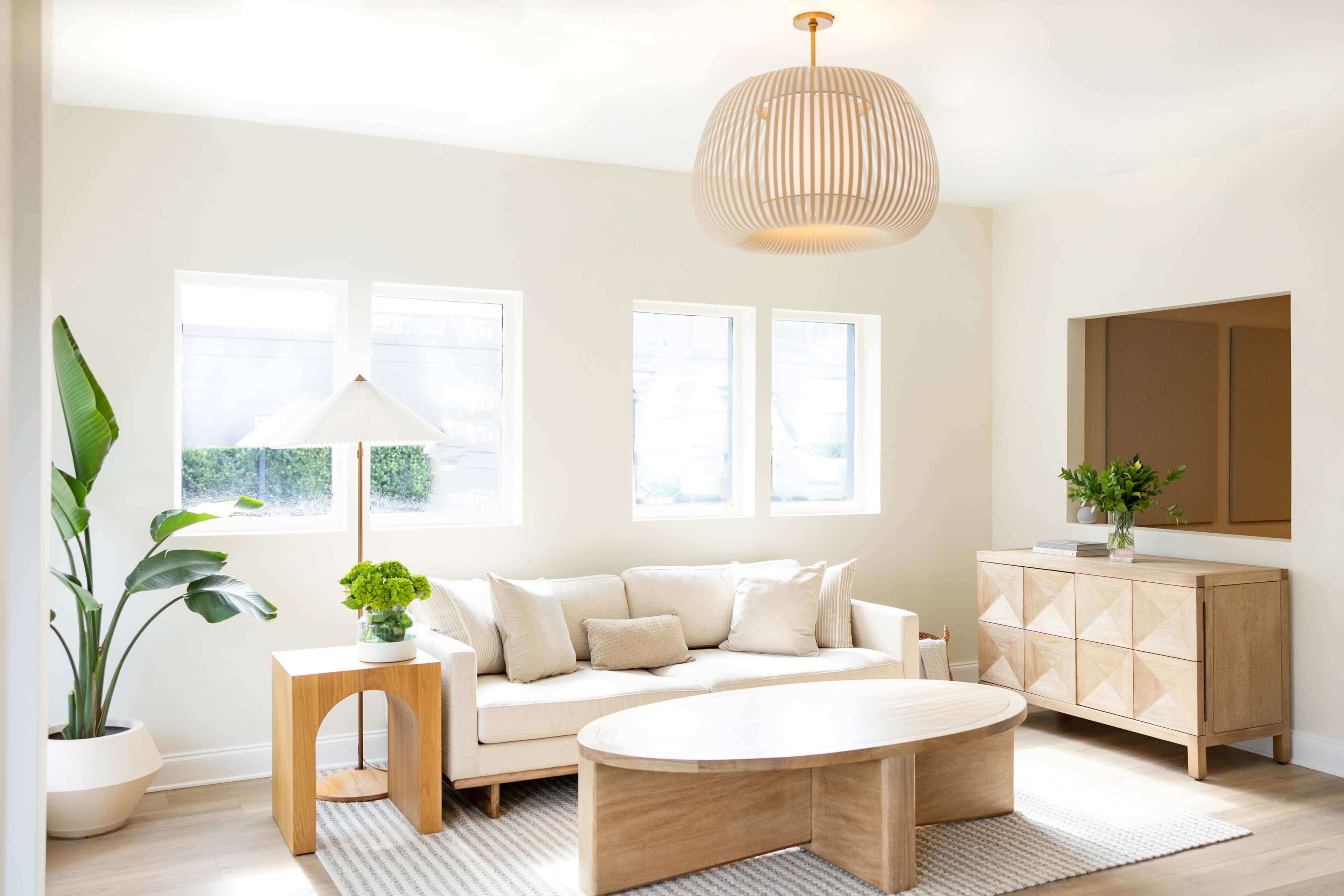 A bright living room featuring a beige sofa, a round coffee table, a decorative plant, and a wooden sideboard under a woven pendant light.