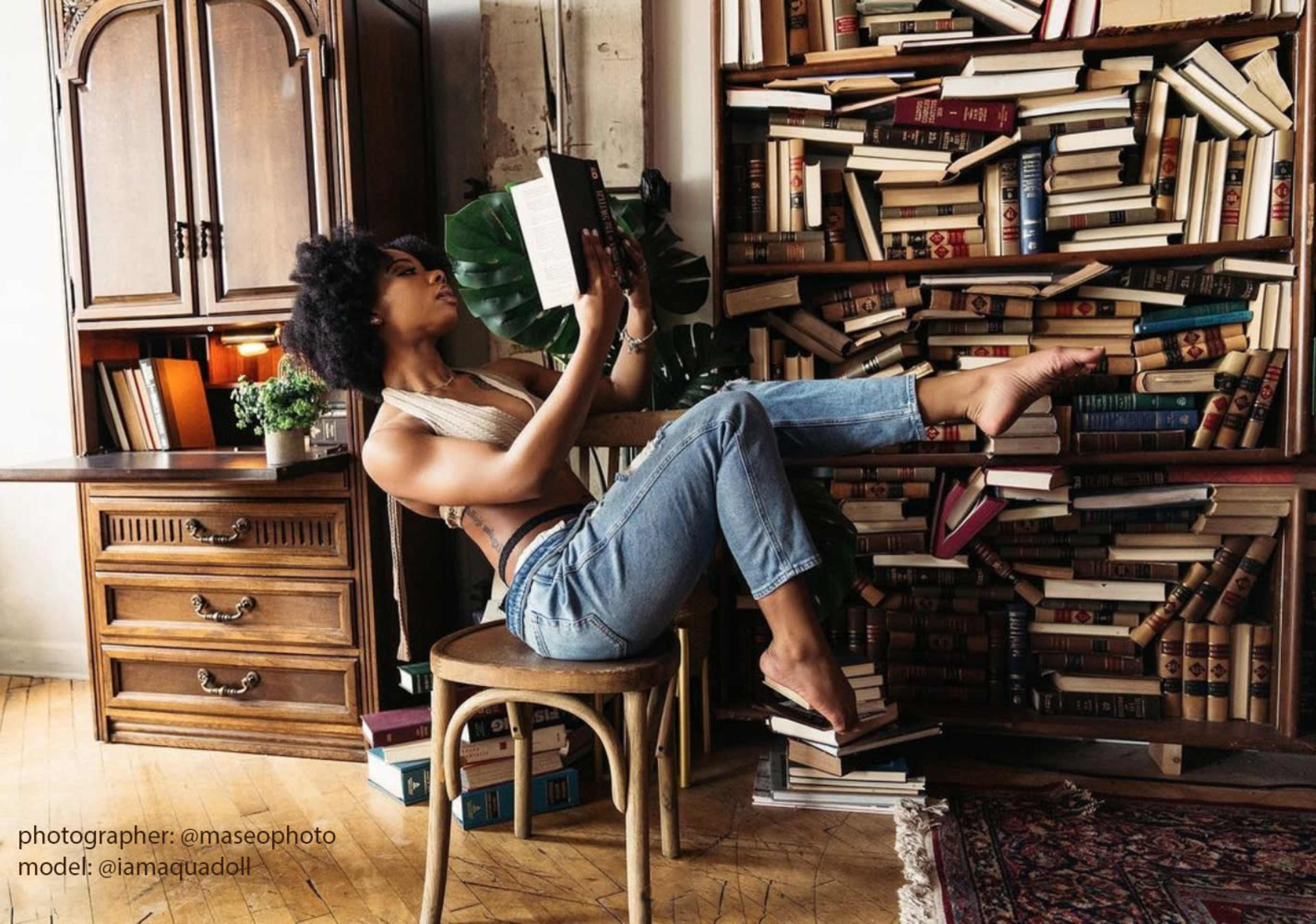A person is seated on a chair, reading a book with one foot resting on a bookshelf surrounded by stacks of books.