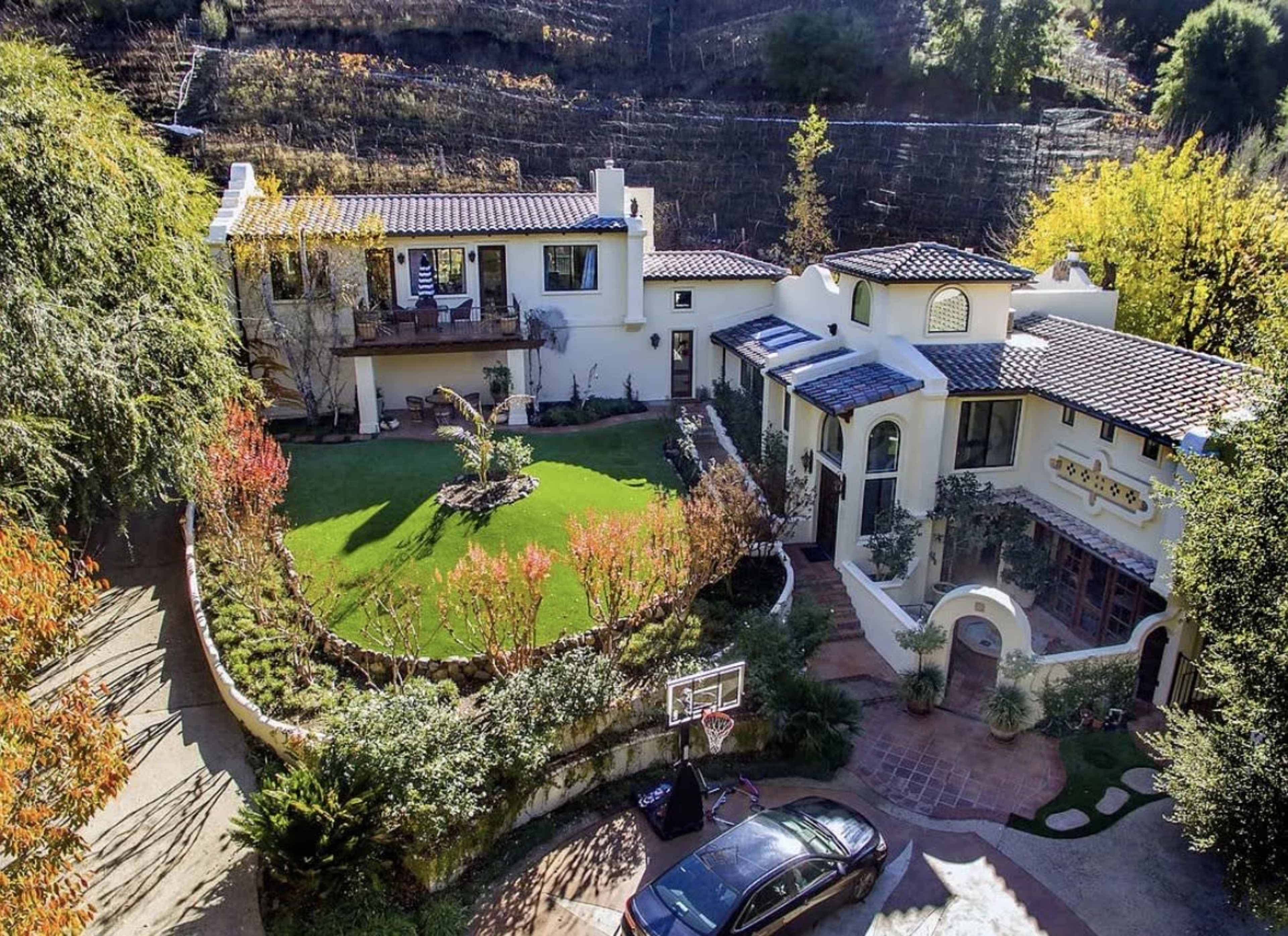 The image shows a large, upscale Mediterranean-style house surrounded by landscaped gardens and greenery, with a basketball hoop in the driveway.
