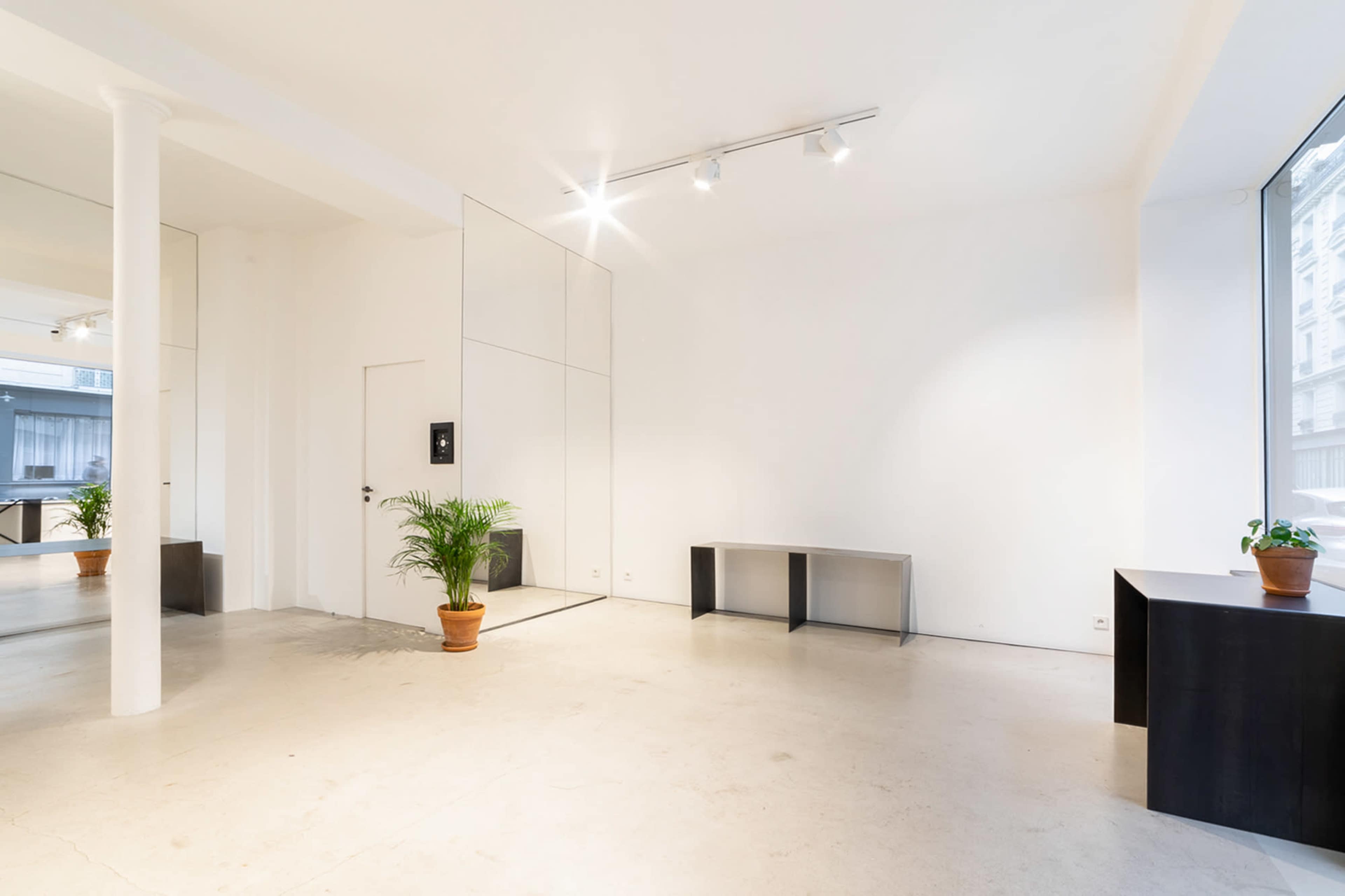 The image shows a spacious, minimalistic interior with white walls, a concrete floor, a large mirror, a potted plant, and simple furniture pieces.