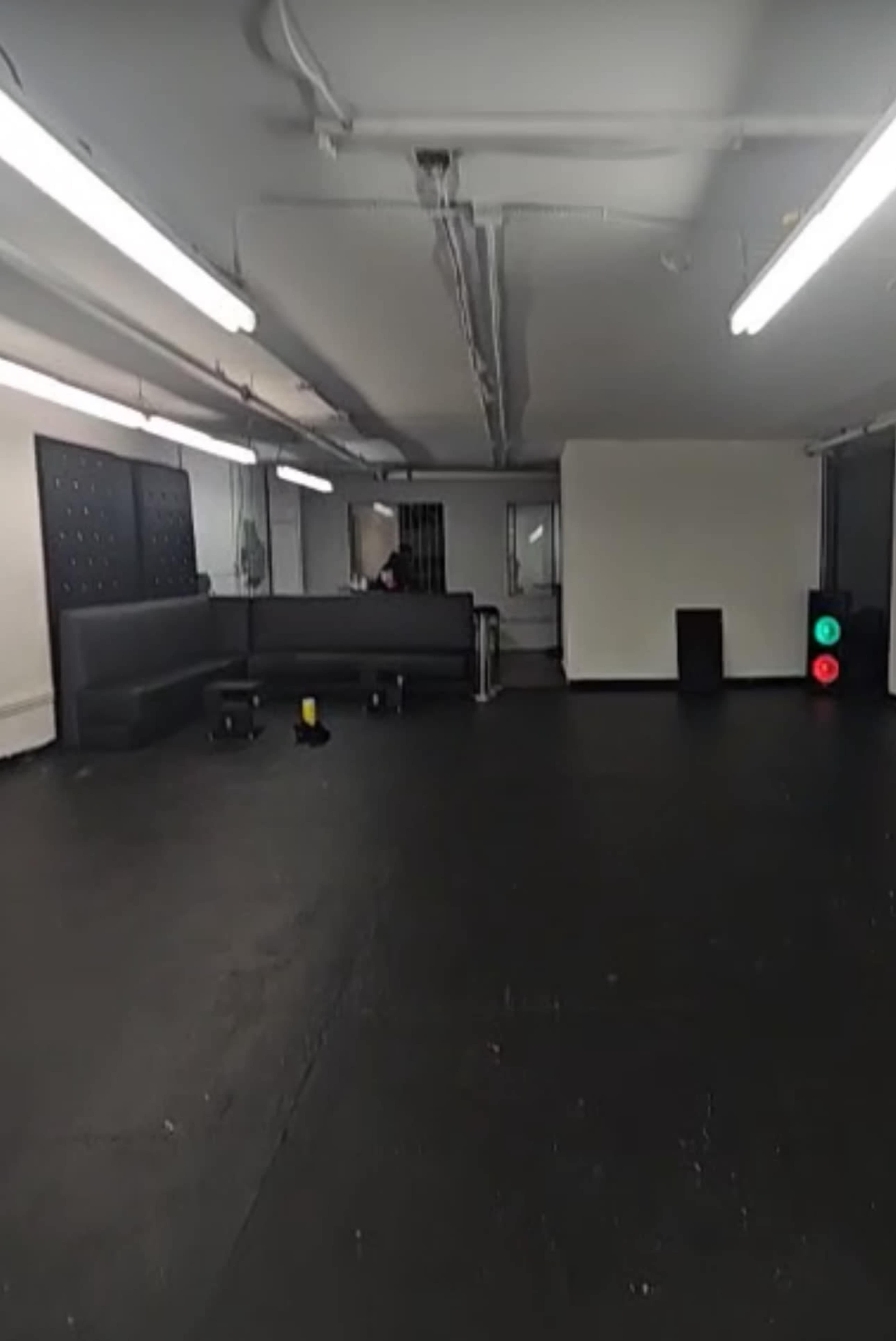 The image shows an empty gym or training space with dark flooring, fluorescent lights, and sparse equipment arranged along the walls.