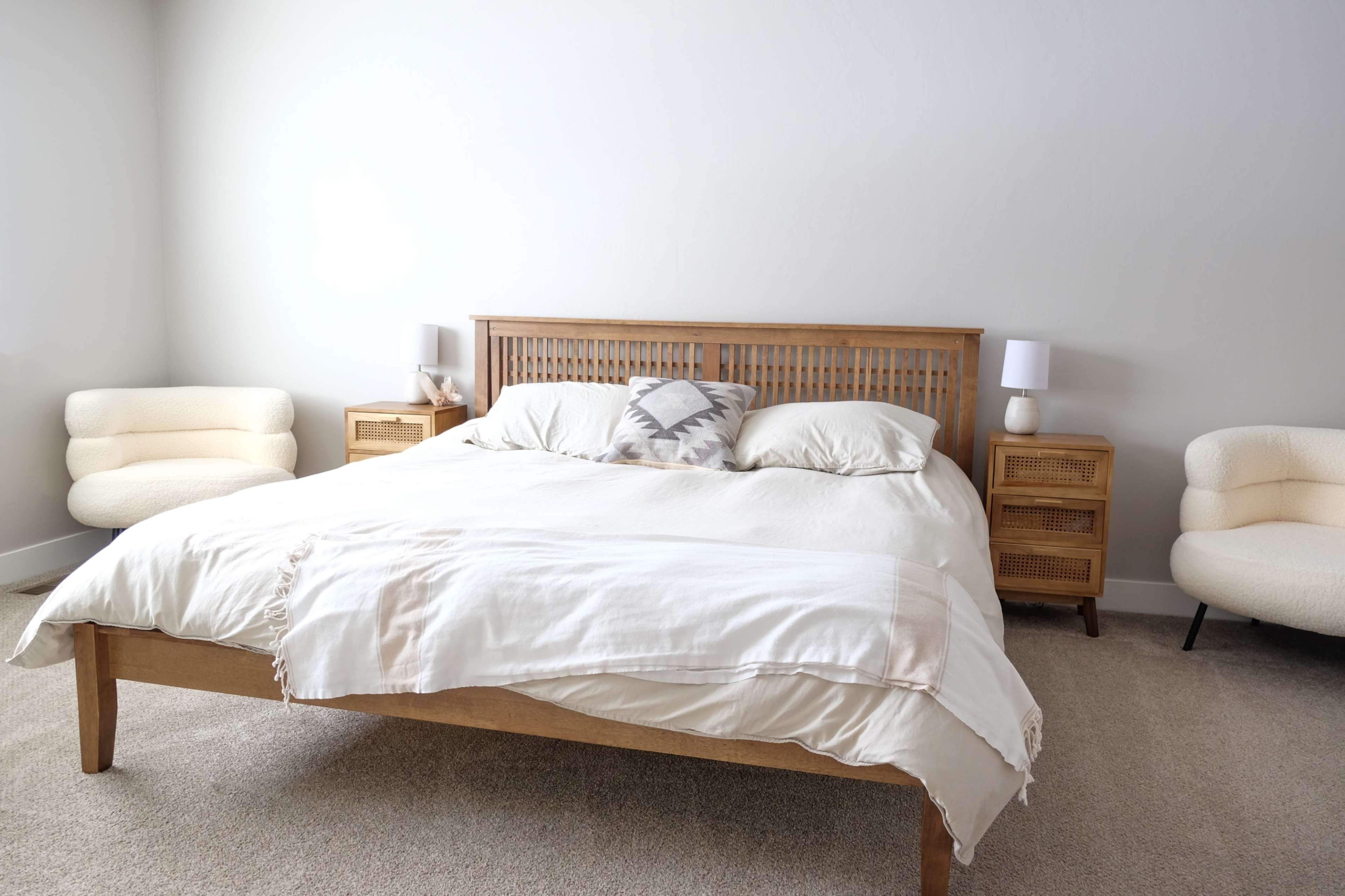 A neatly arranged bedroom features a wooden bed with a light-colored comforter and two bedside tables, each with a lamp, against a plain wall.