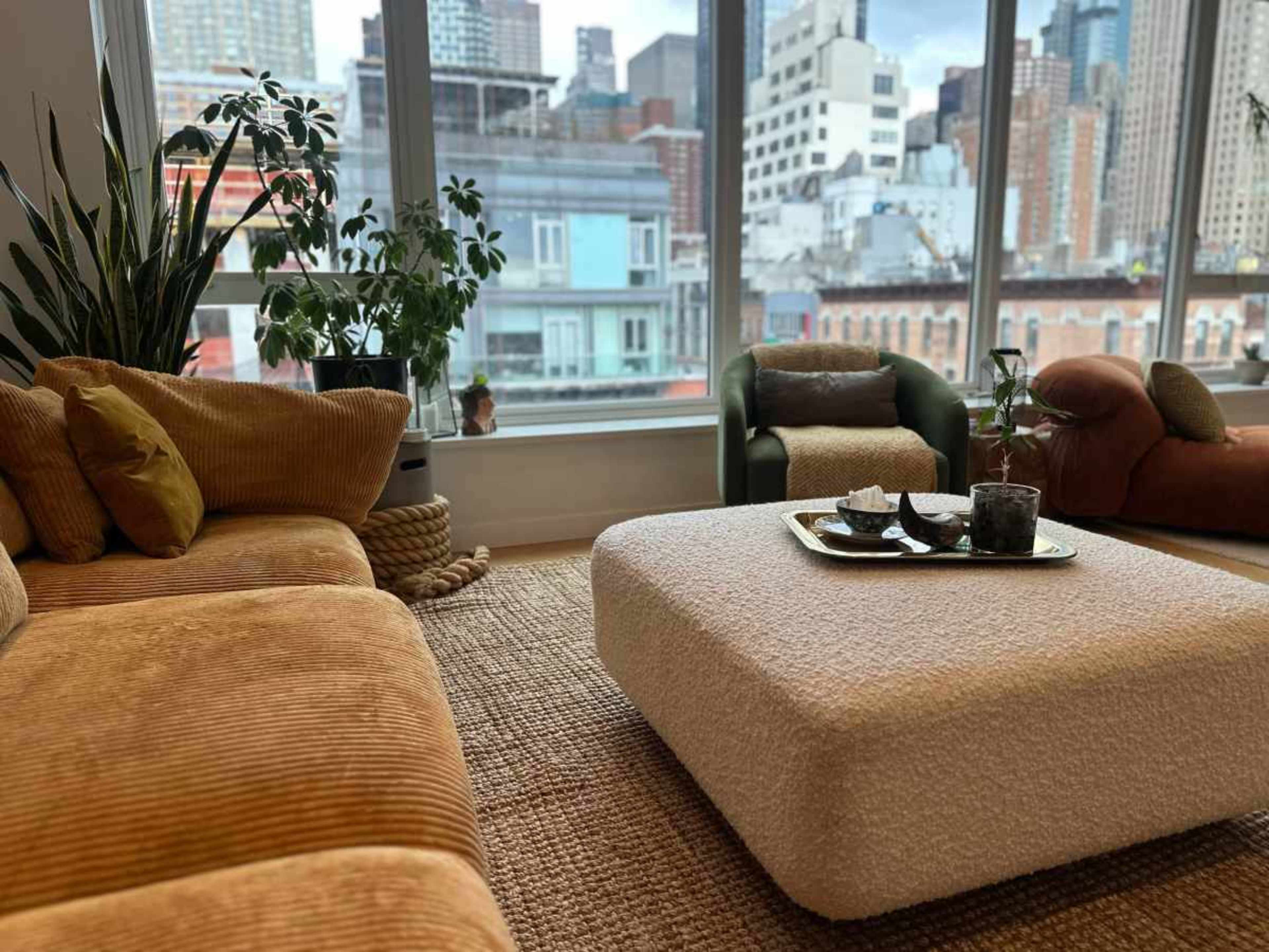 The scene features a cozy living room with two sofas, a textured coffee table, and large windows revealing a city skyline.