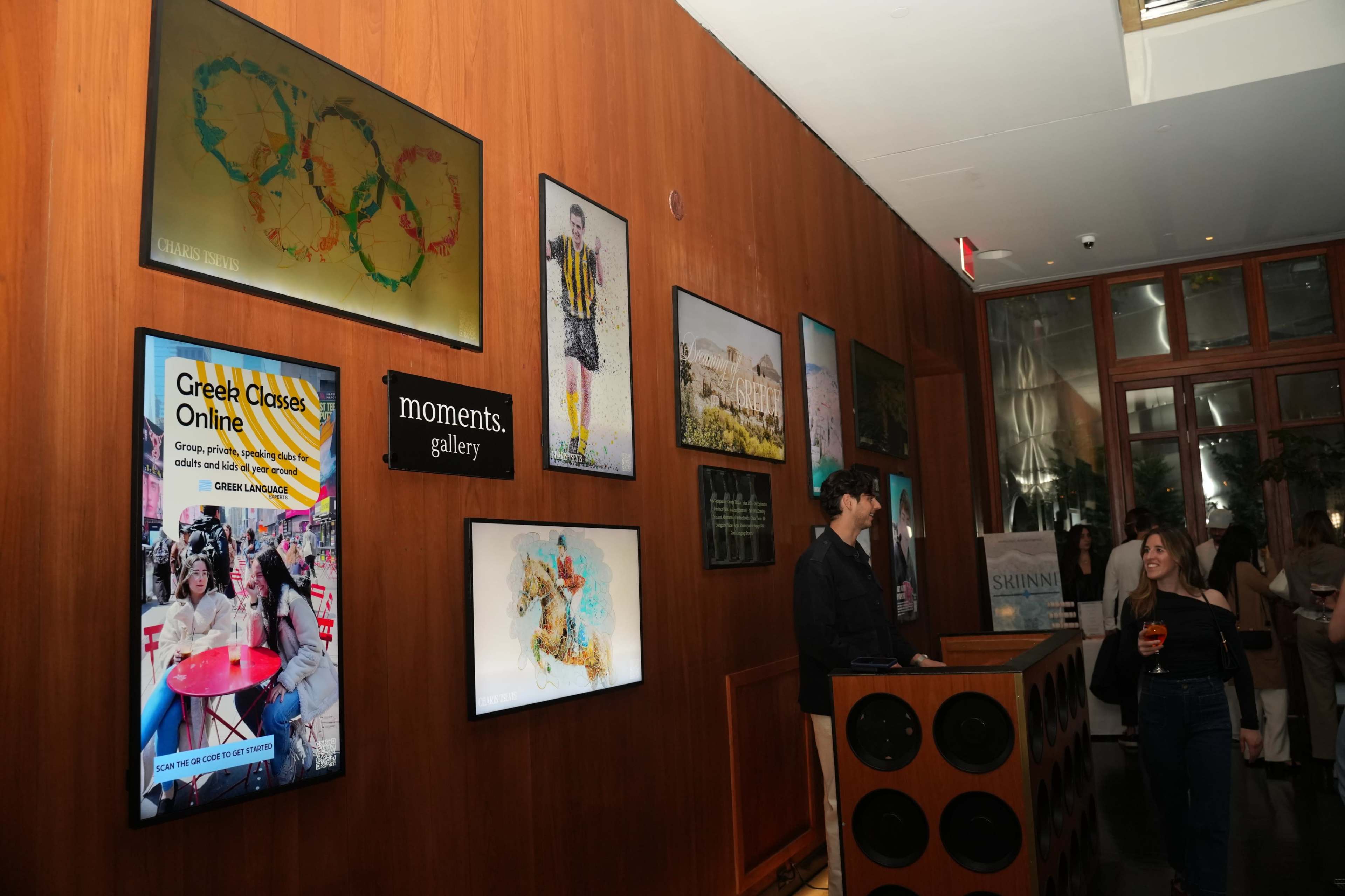 The image shows a gallery wall with various framed artworks and a reception area where a staff member interacts with a visitor.