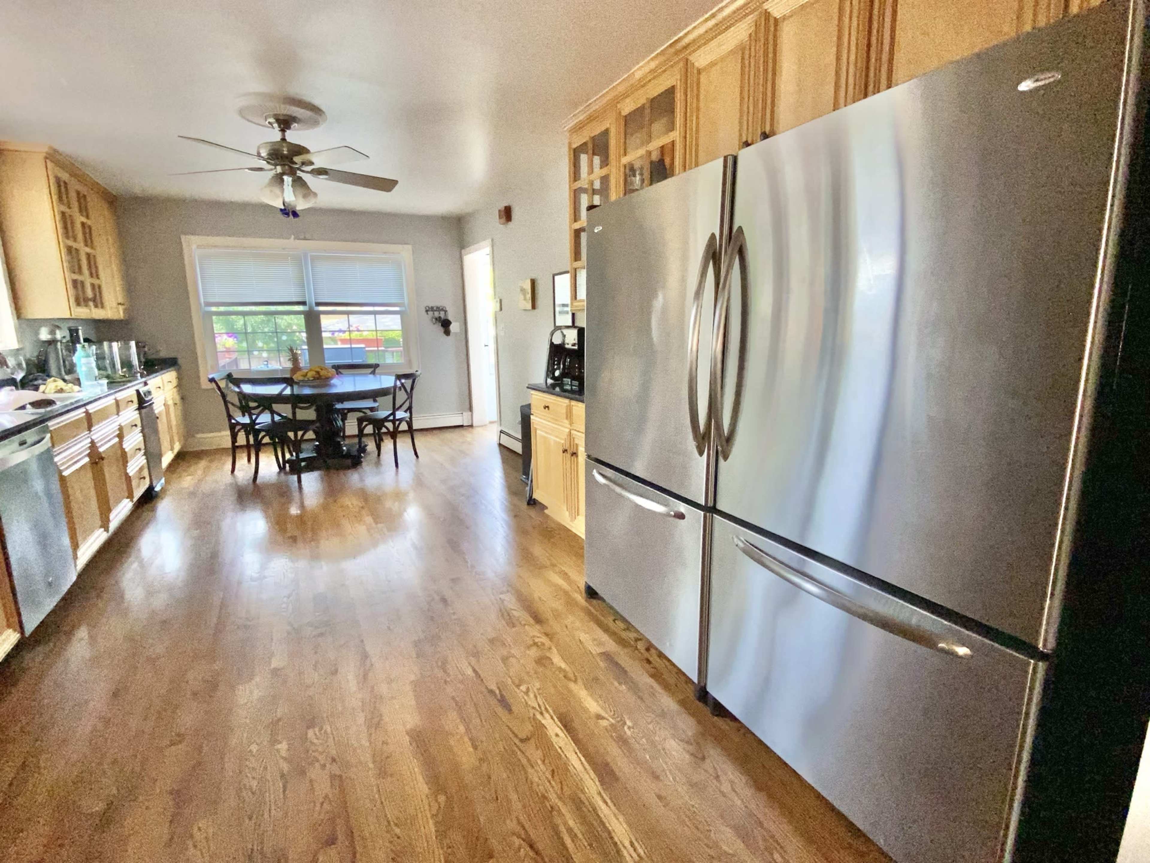 The image shows a modern kitchen with wooden cabinets, a large stainless steel refrigerator, and a dining table set for six.