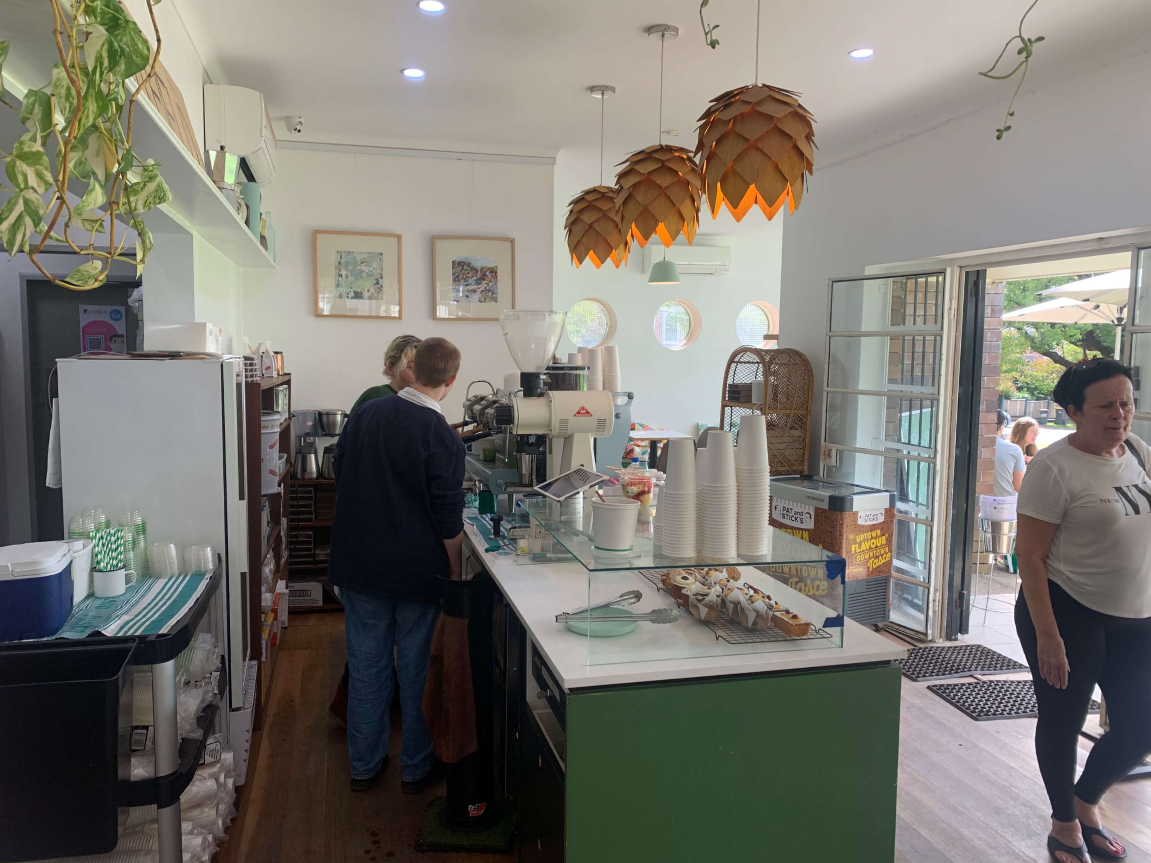 A coffee shop interior features two baristas behind the counter, surrounded by various coffee-making equipment and display items, with patrons visible outside through an open door.