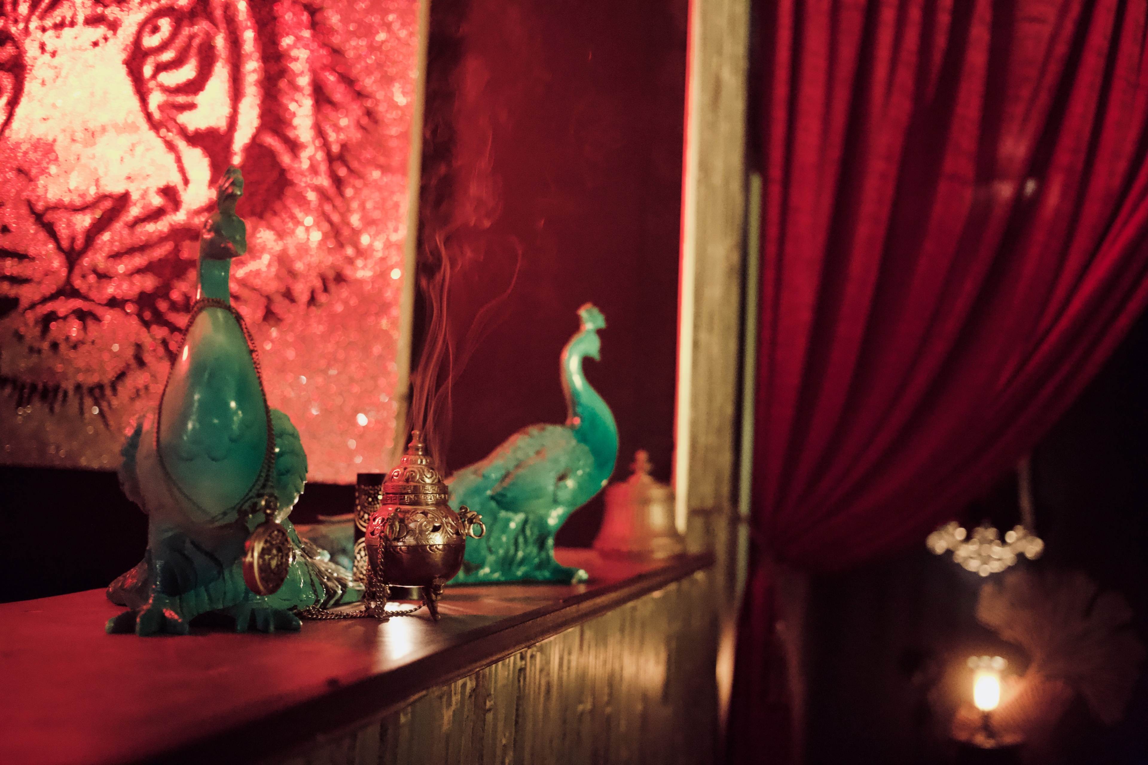 Two blue glass peacock statues and a small ornate incense holder on a wooden ledge, with red curtains hanging in the background.