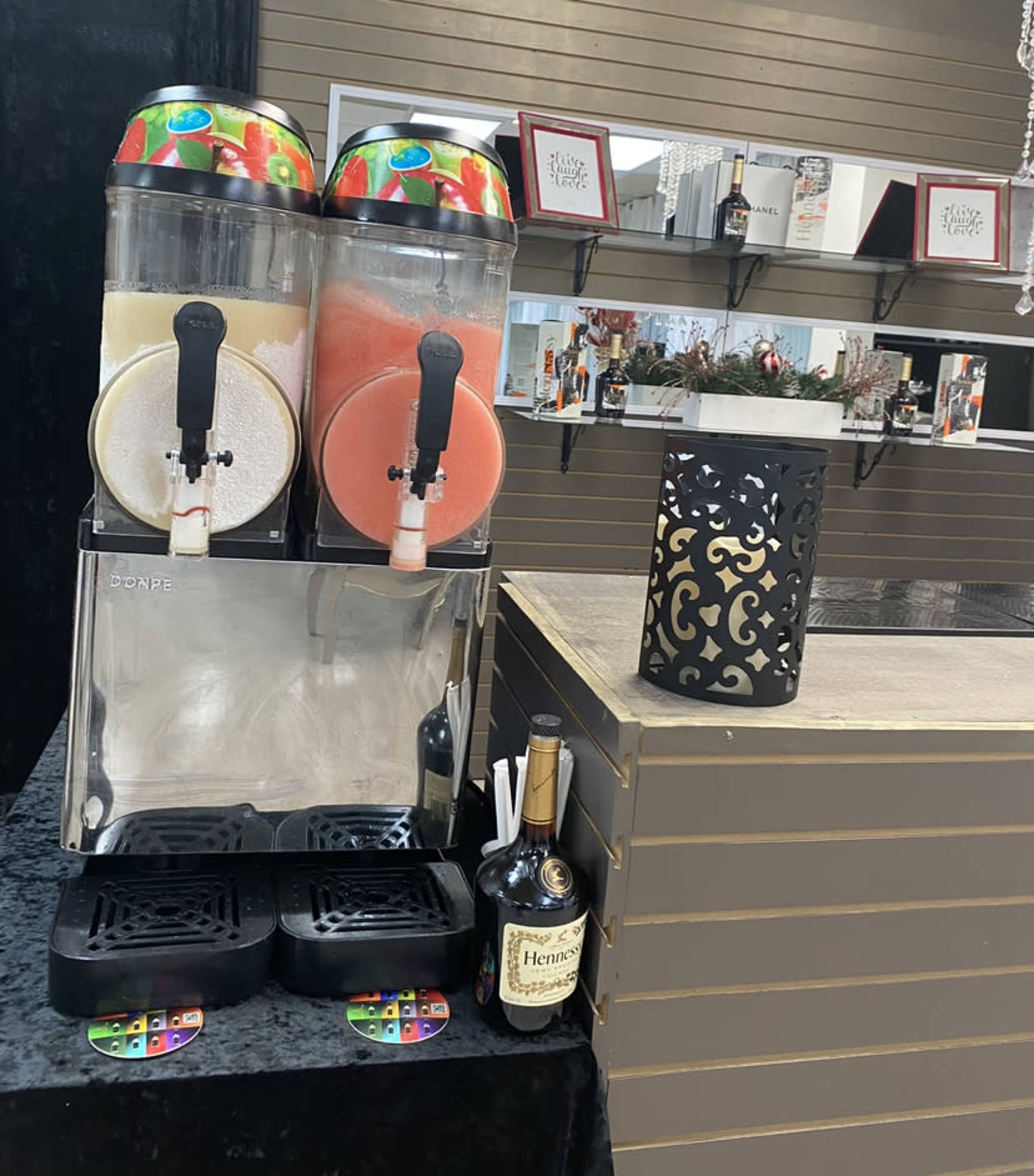 The image shows a beverage dispenser with two slushy machines, one filled with a yellow drink and the other with a red drink, beside a decorative lantern and a bottle of Hennessy.
