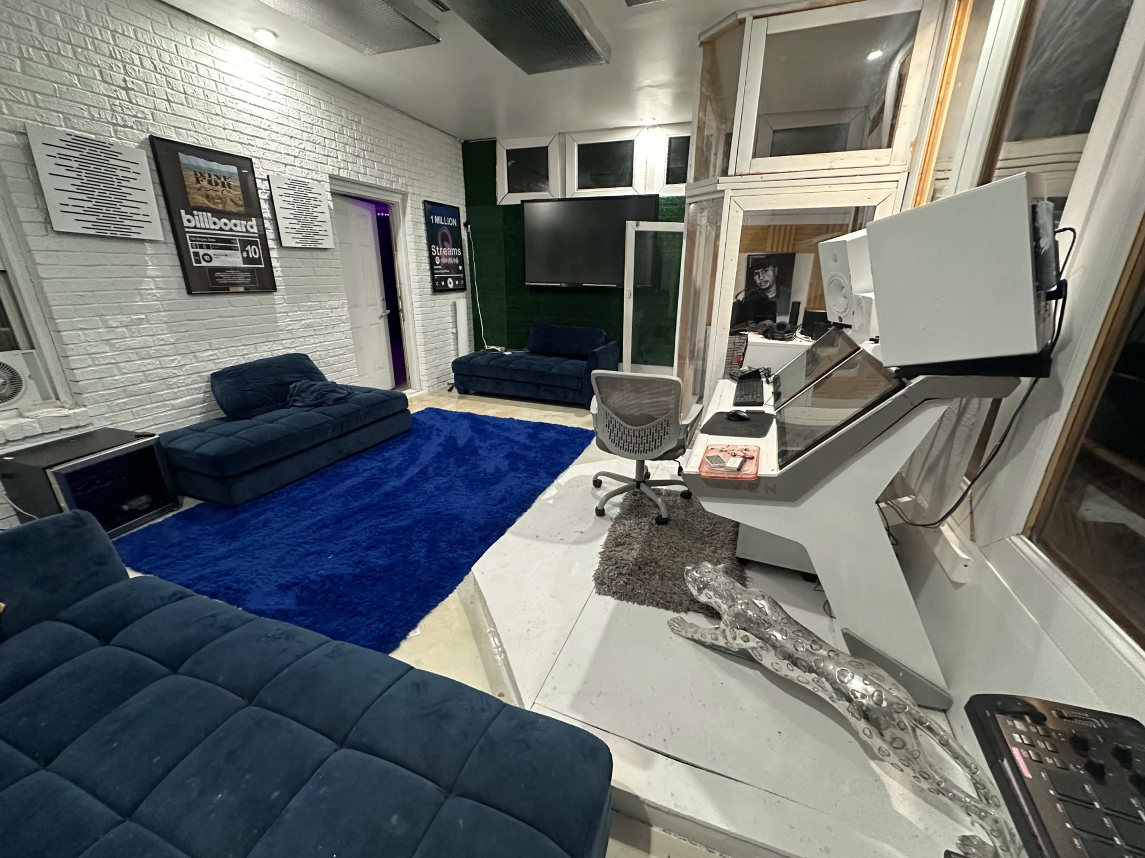 The image depicts a modern recording studio with blue couches, a plush rug, and a workstation equipped with audio equipment and a computer.