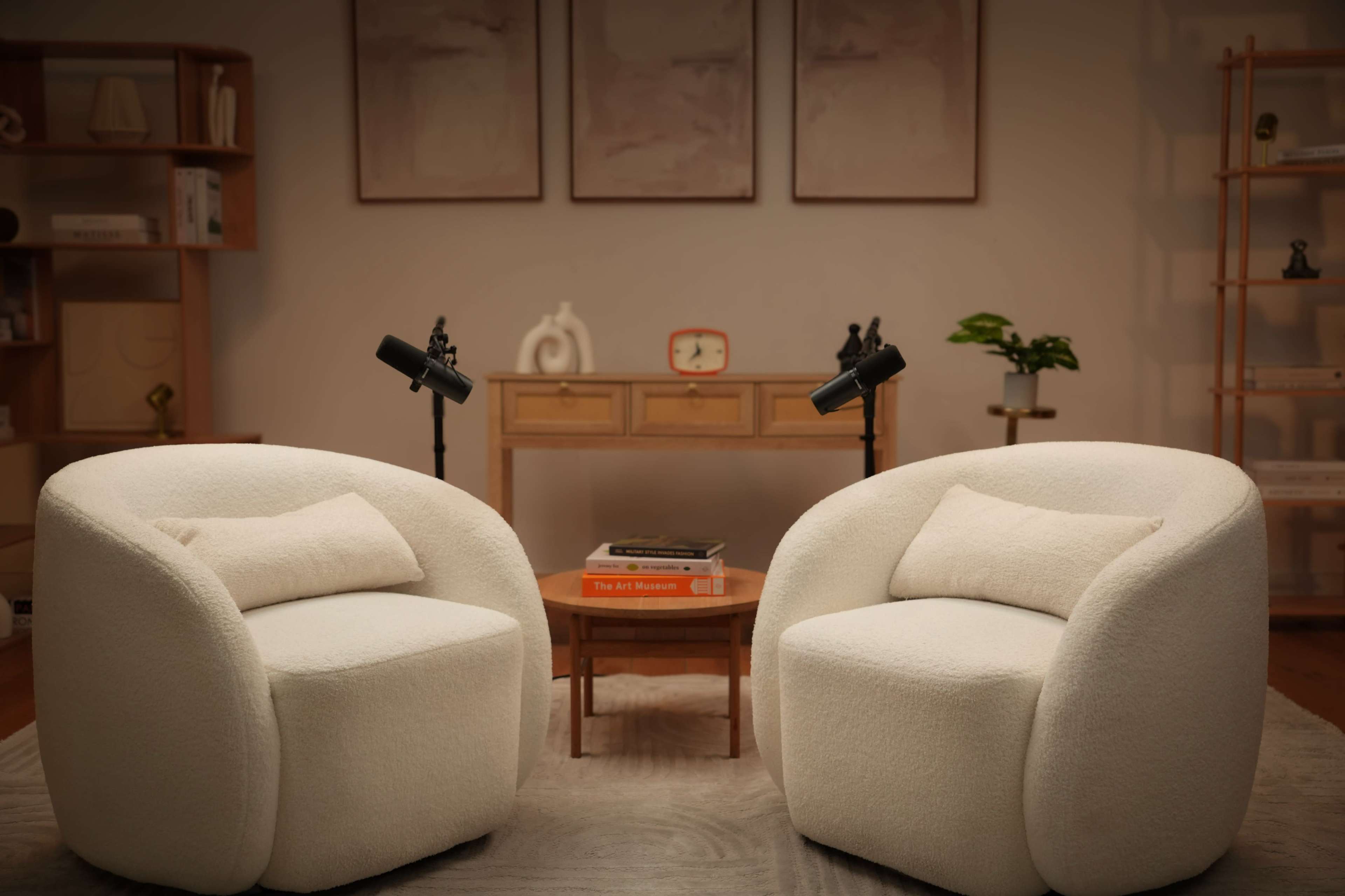 The image shows two white, rounded armchairs facing each other on a rug, with a small wooden table and a decorative shelf in the background.
