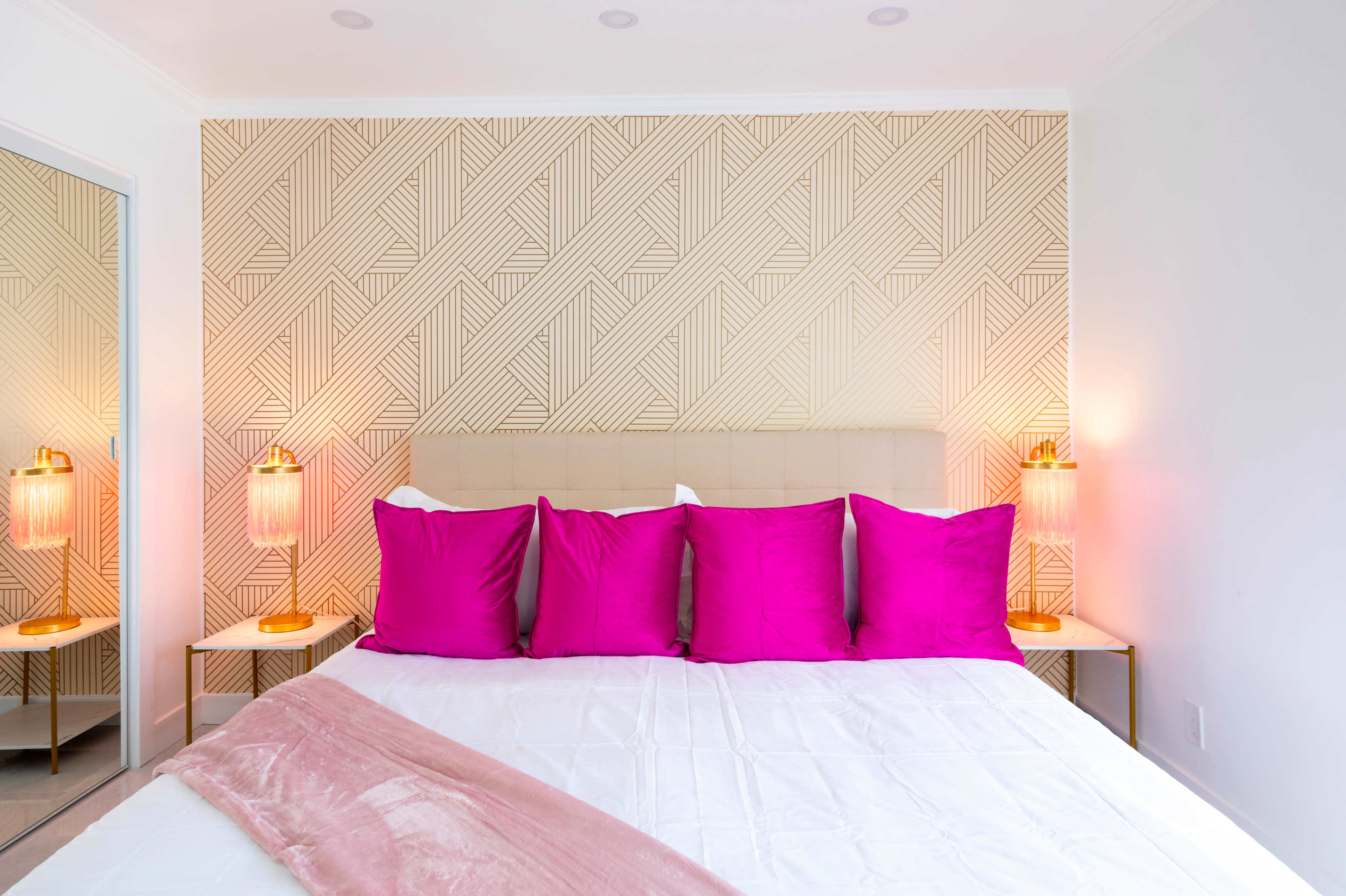 The image shows a neatly arranged bedroom featuring a bed with a beige headboard, adorned with five pink pillows, against a patterned wall, with two bedside lamps on either side.