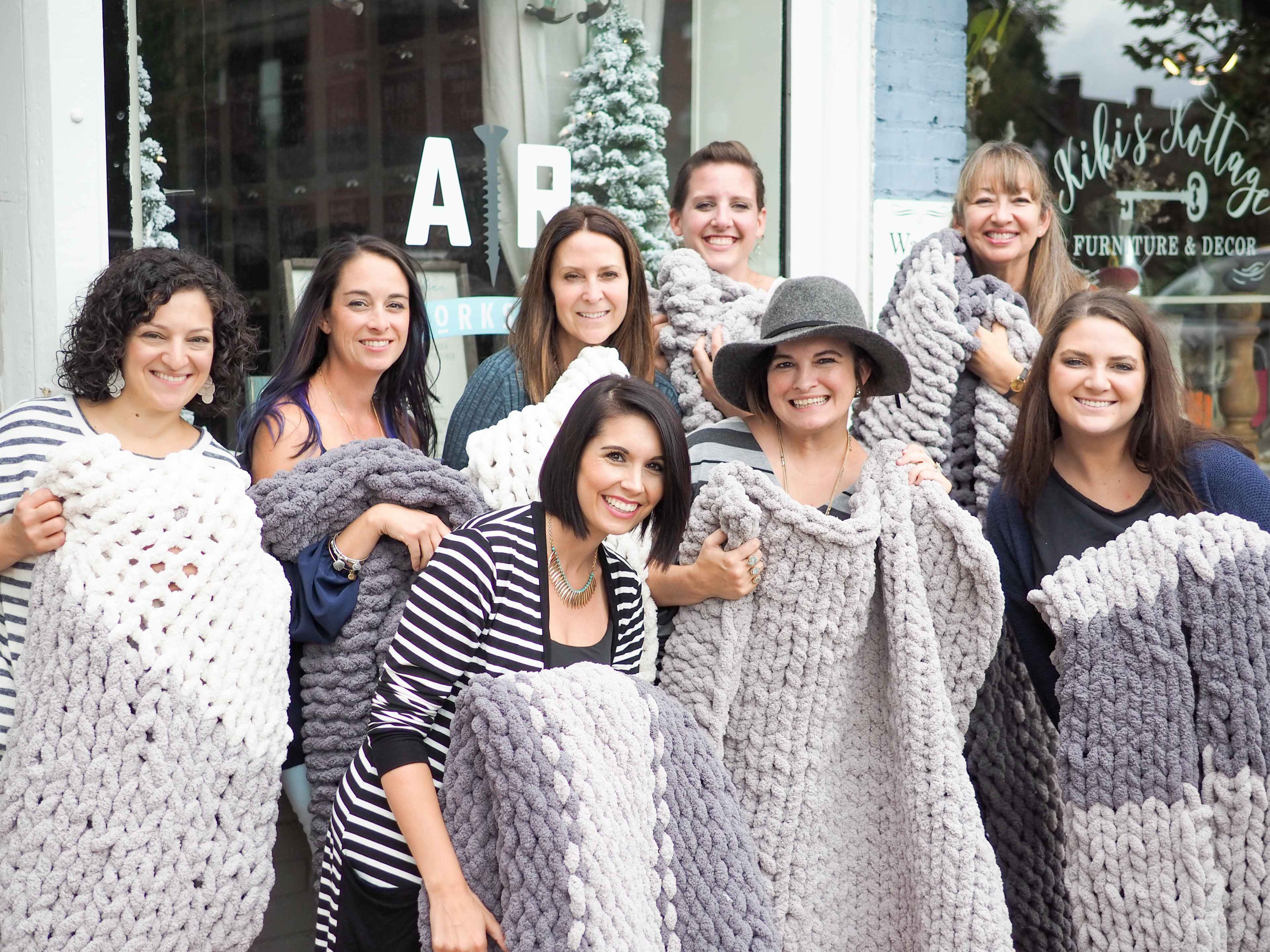 A group of nine women hold large knitted blankets outside a store with a decorated window.