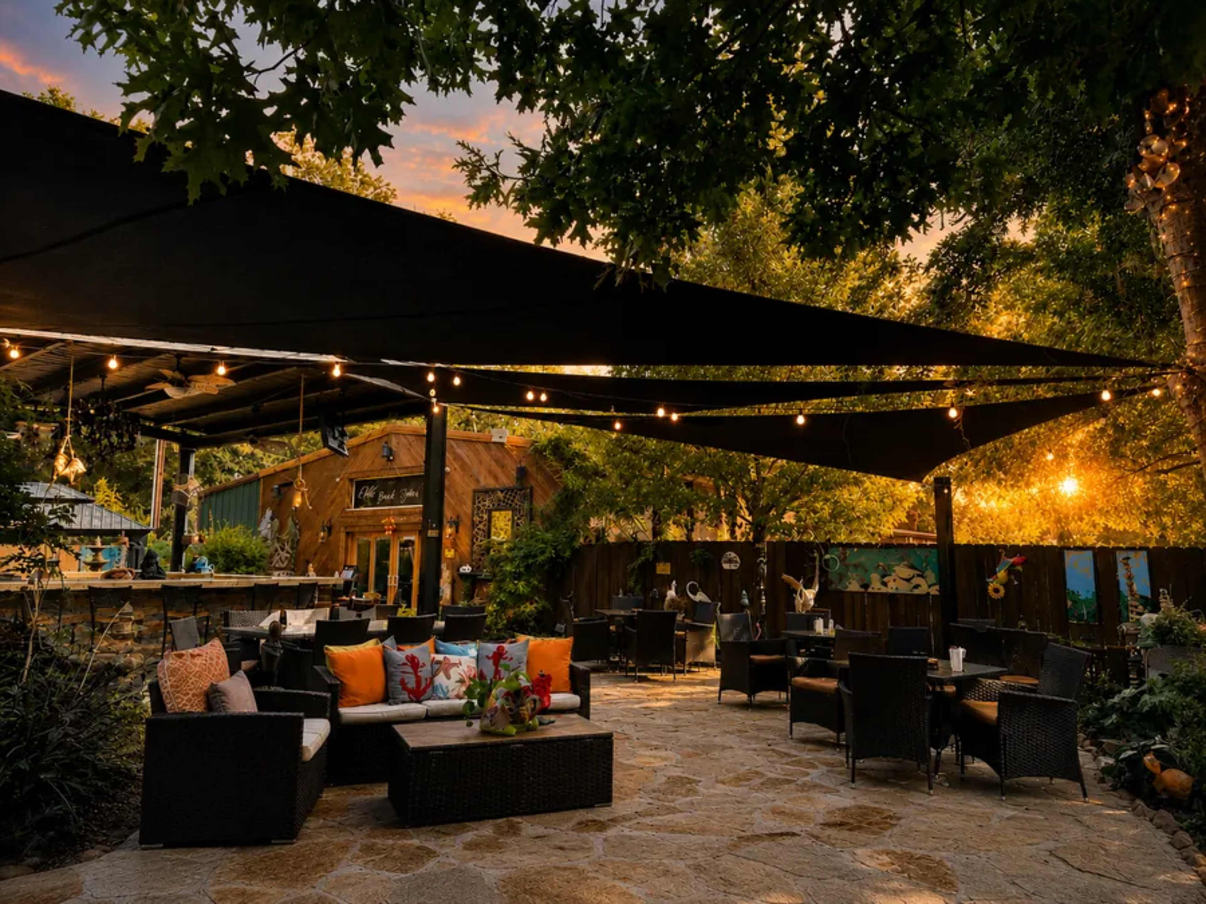 The image shows a patio area with outdoor furniture, shaded by large canopies, surrounded by greenery and illuminated by string lights during sunset.