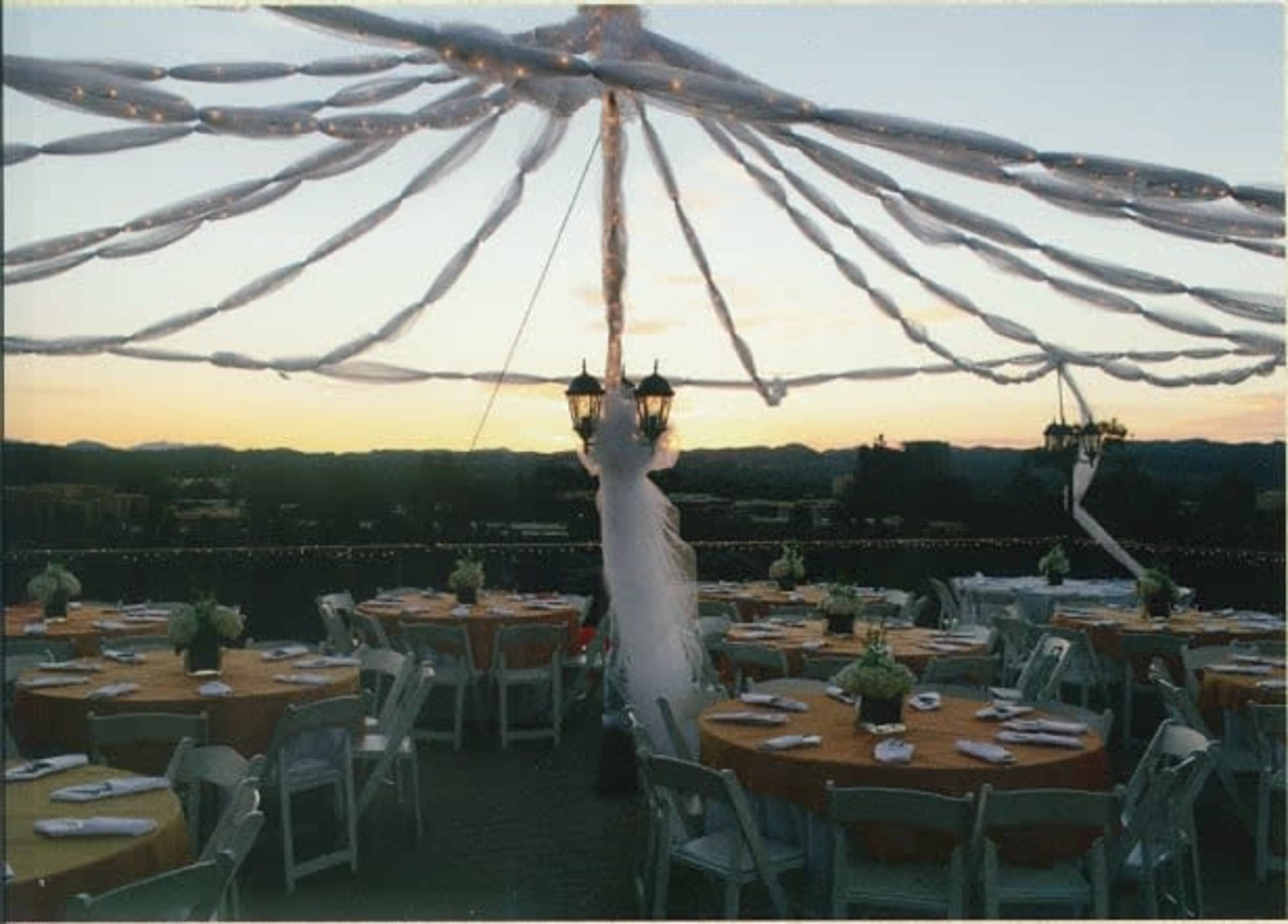 The image shows a decorated outdoor event space with several tables covered in orange tablecloths, under a canopy of white drapes and string lights at sunset.