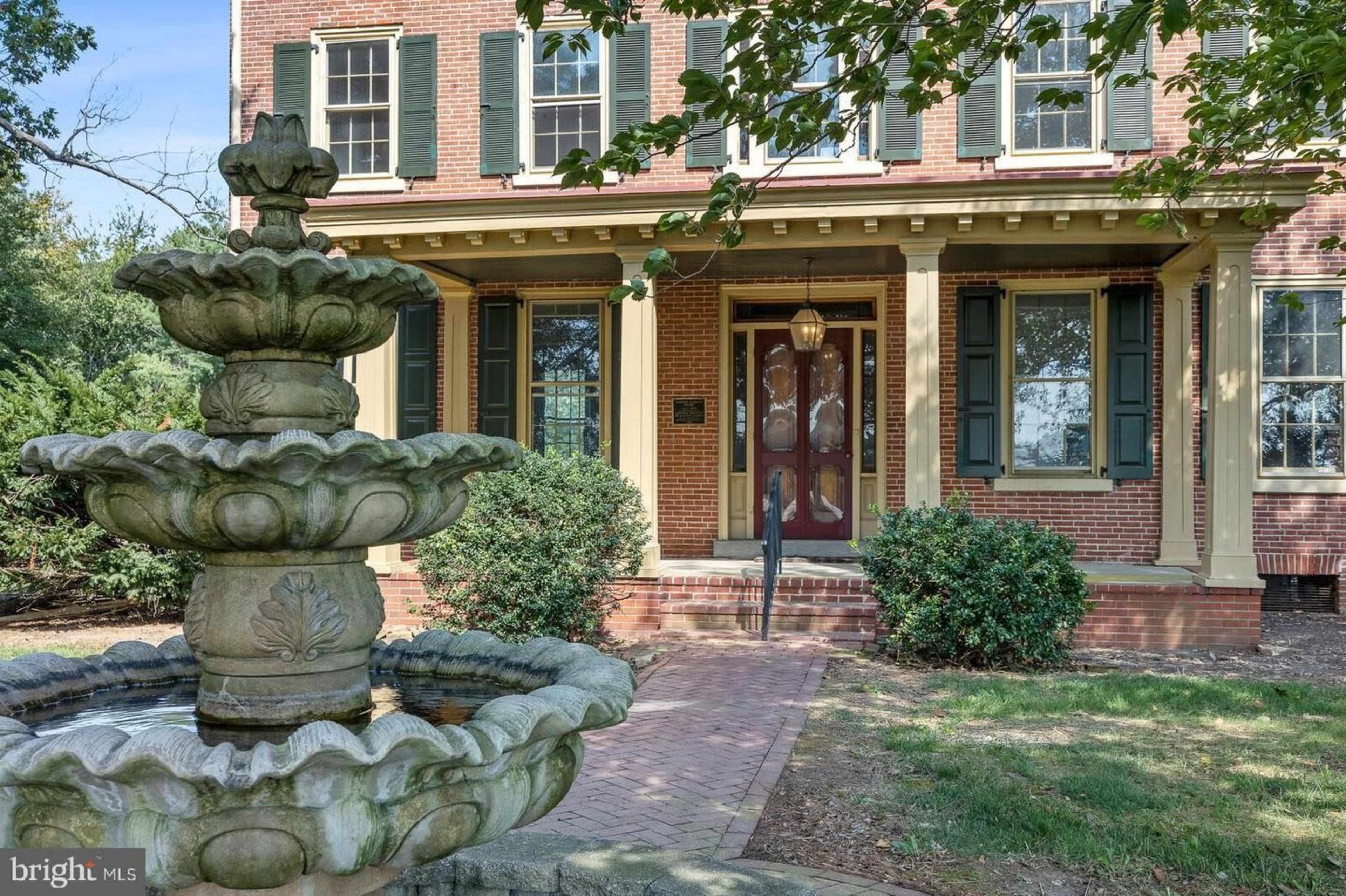 A brick house features a front porch with green shutters and a three-tiered fountain in the foreground.