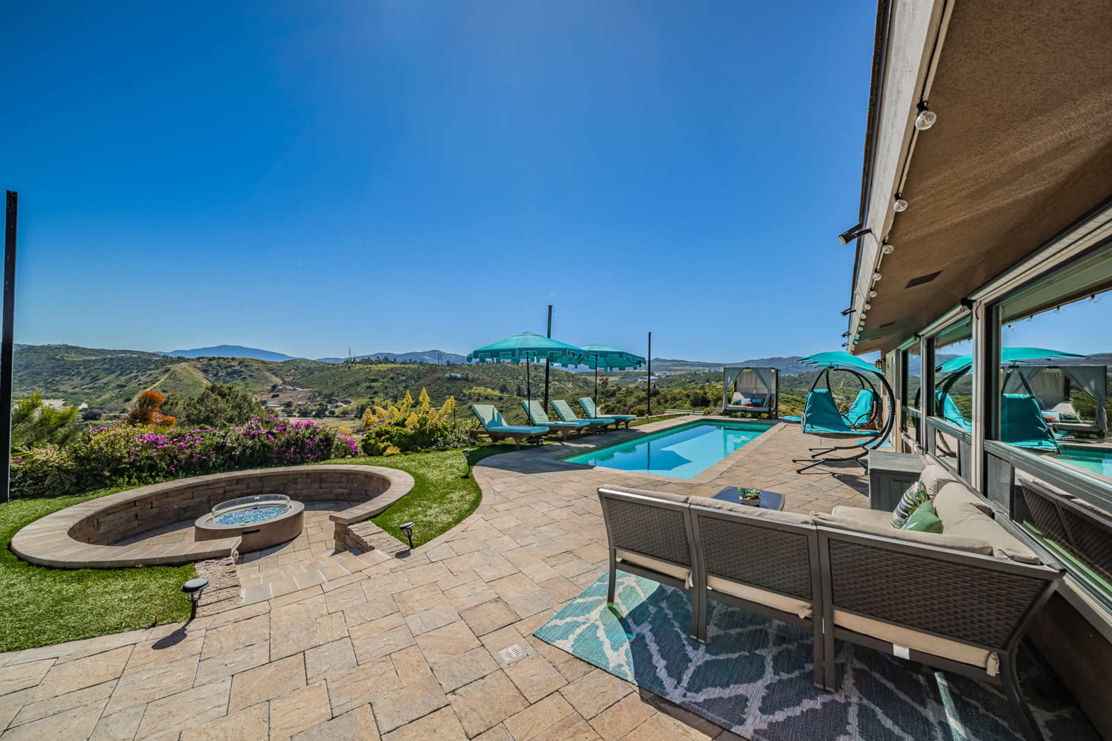A backyard pool area with lounge chairs, a hot tub, and scenic hills in the background under a clear blue sky.