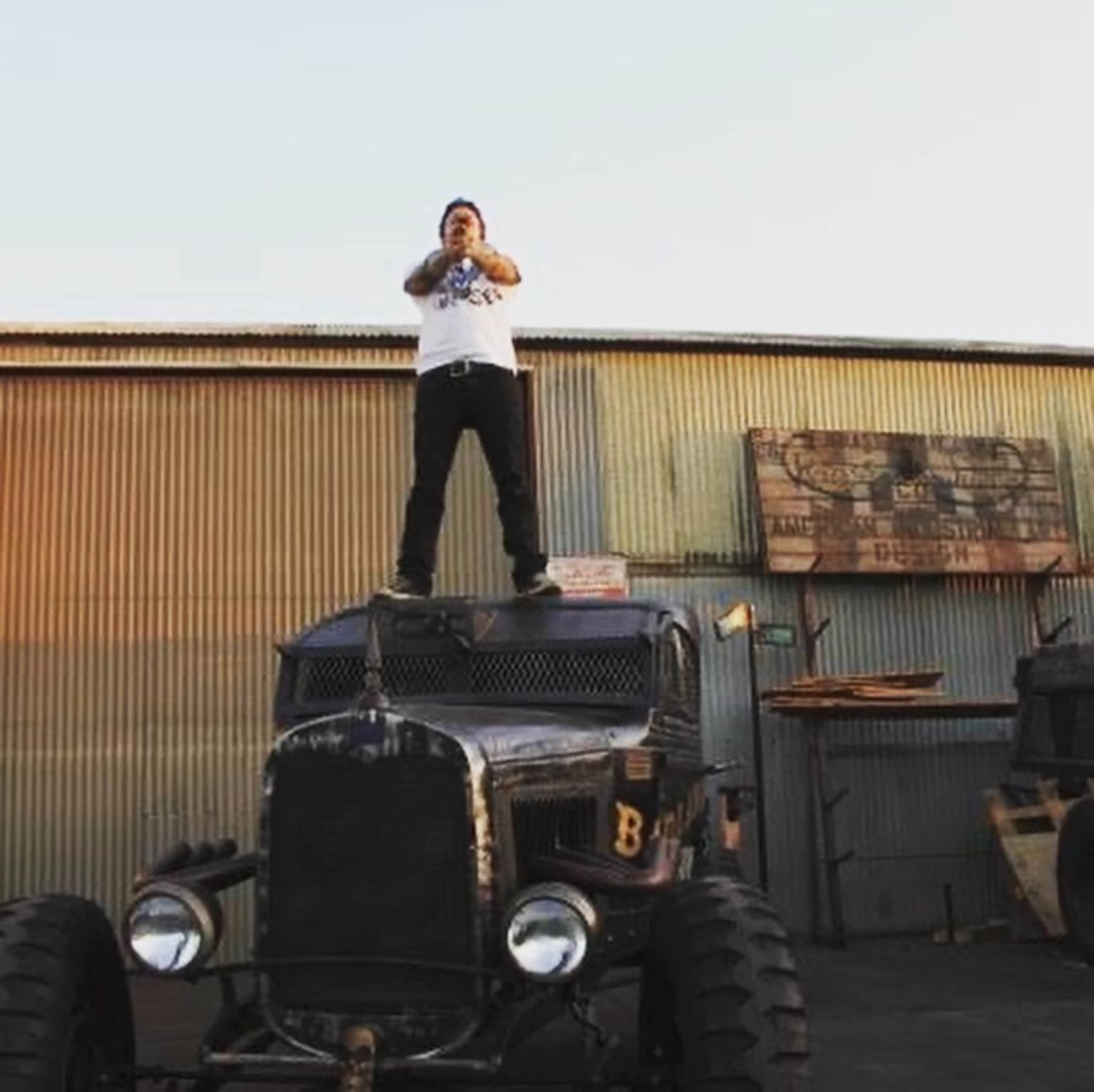 A person stands on the roof of an antique truck in front of a corrugated metal building.