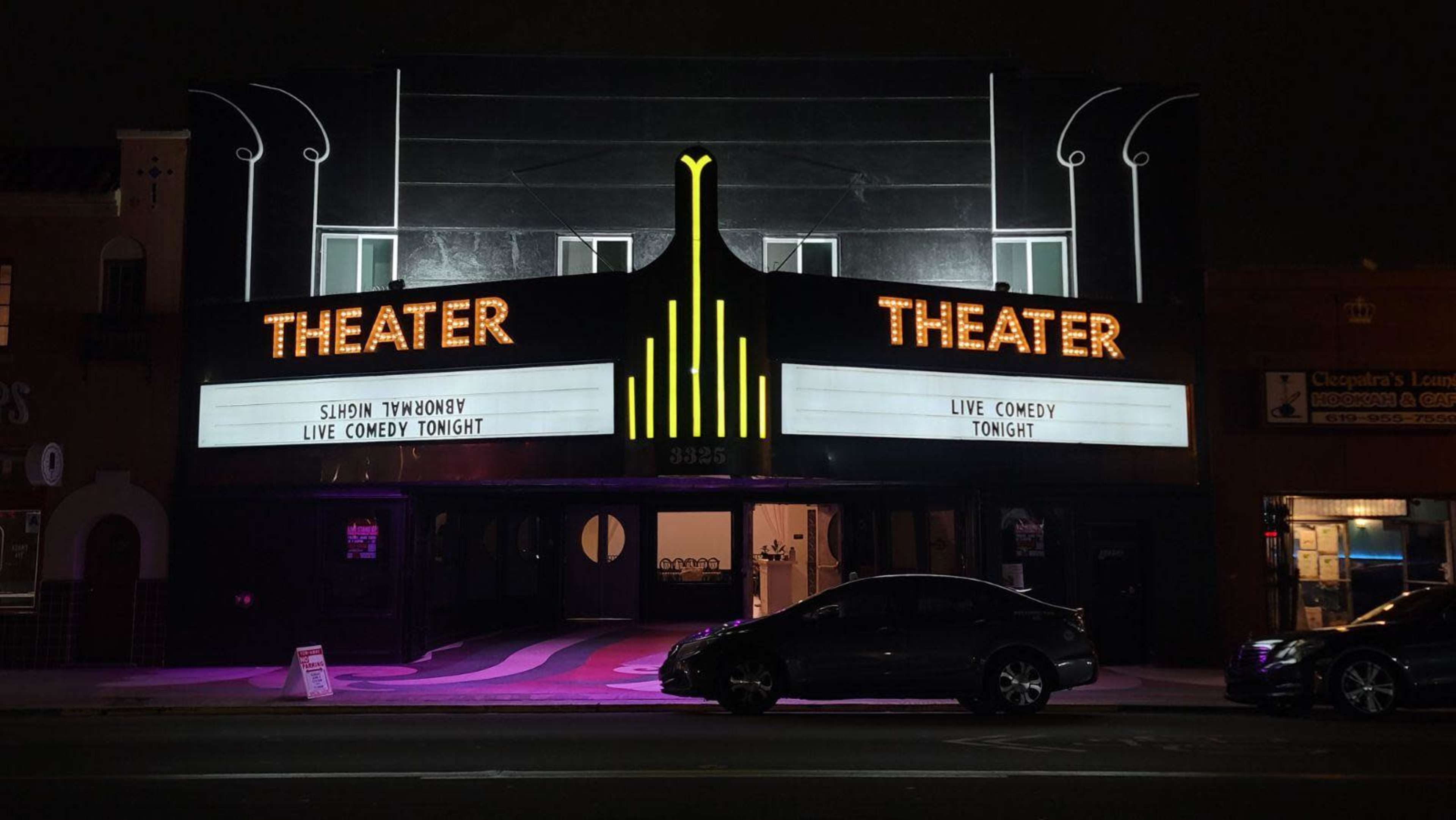 A theater building displays a marquee announcing a live comedy event, with a sleek, illuminated facade and a parked car in front.