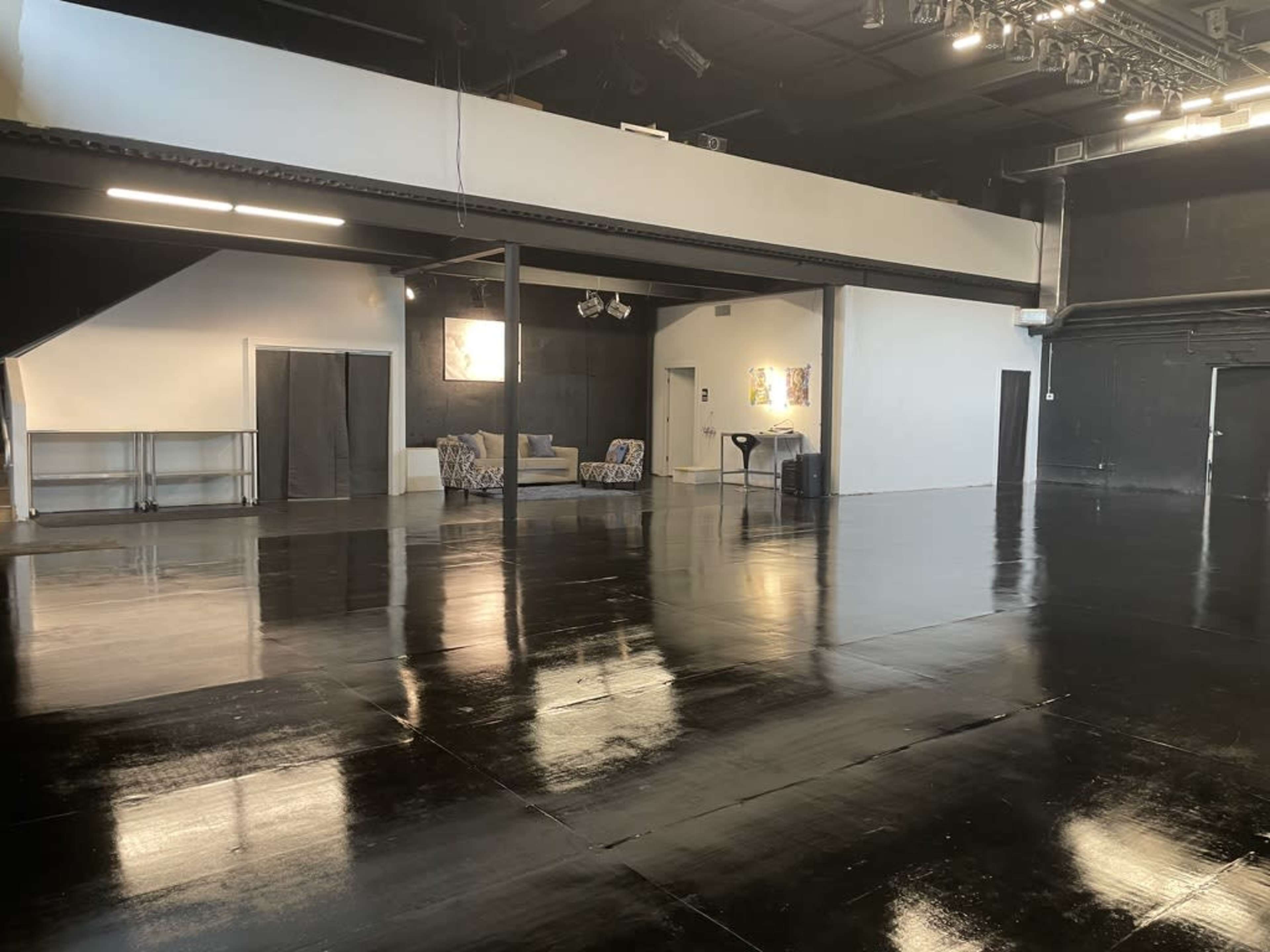 The image shows a spacious, polished studio with black flooring, featuring a seating area and two doors in the background.