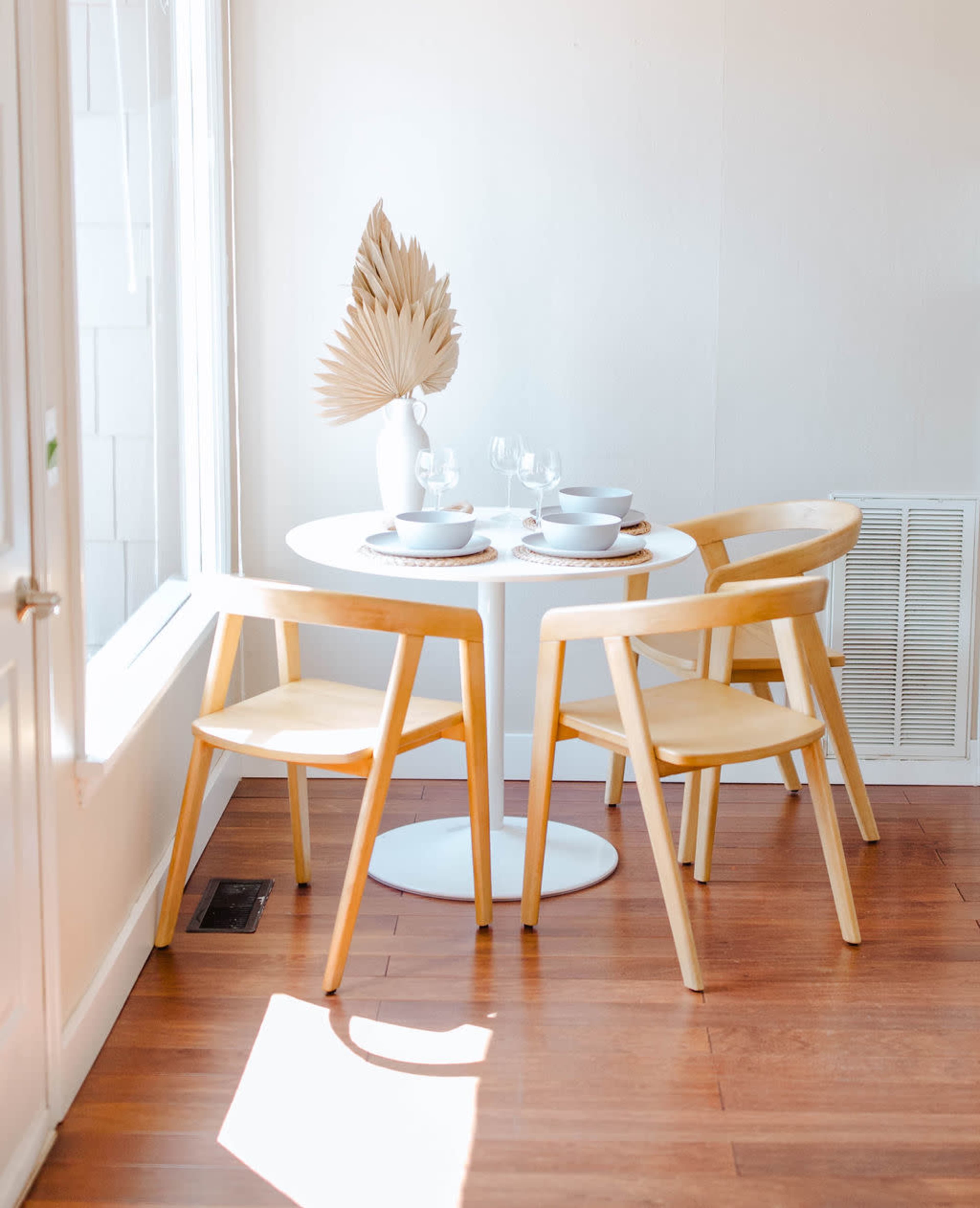 A small dining table set with plates and glasses is surrounded by two wooden chairs in a bright, minimalist room.