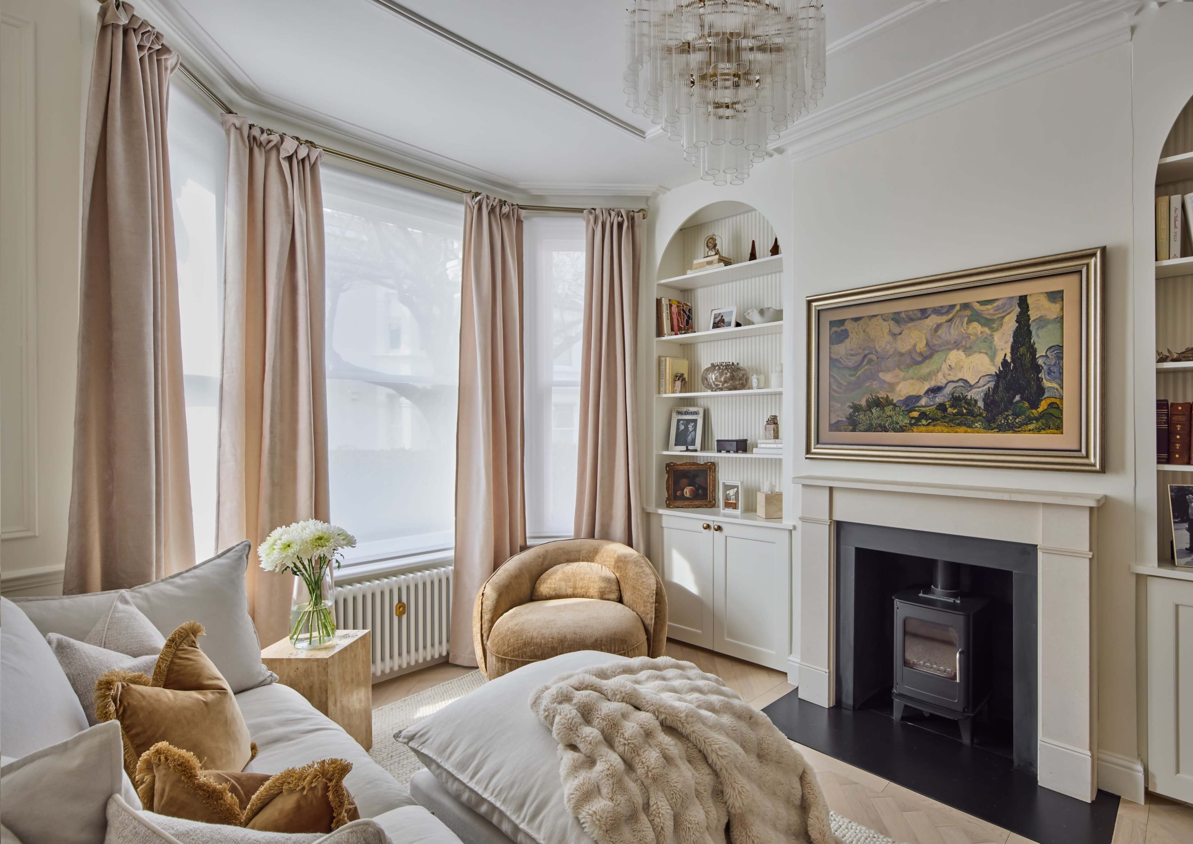 The image shows a cozy living room featuring a comfortable sofa, a round chair, a fireplace, built-in shelves with decorative items, and large windows dressed with long curtains.