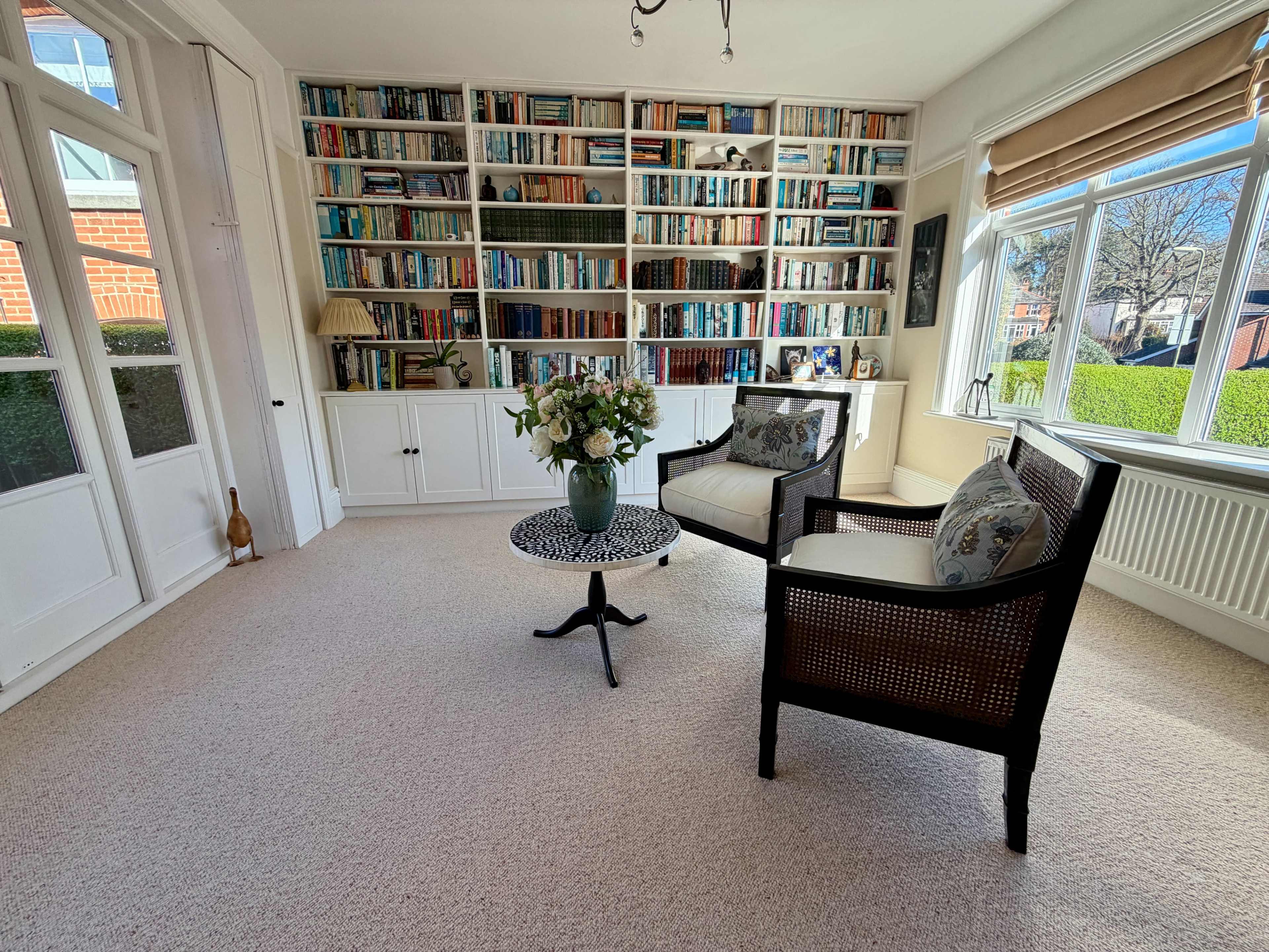 The image shows a well-lit room with two chairs and a small table, set against a wall lined with bookshelves filled with books.