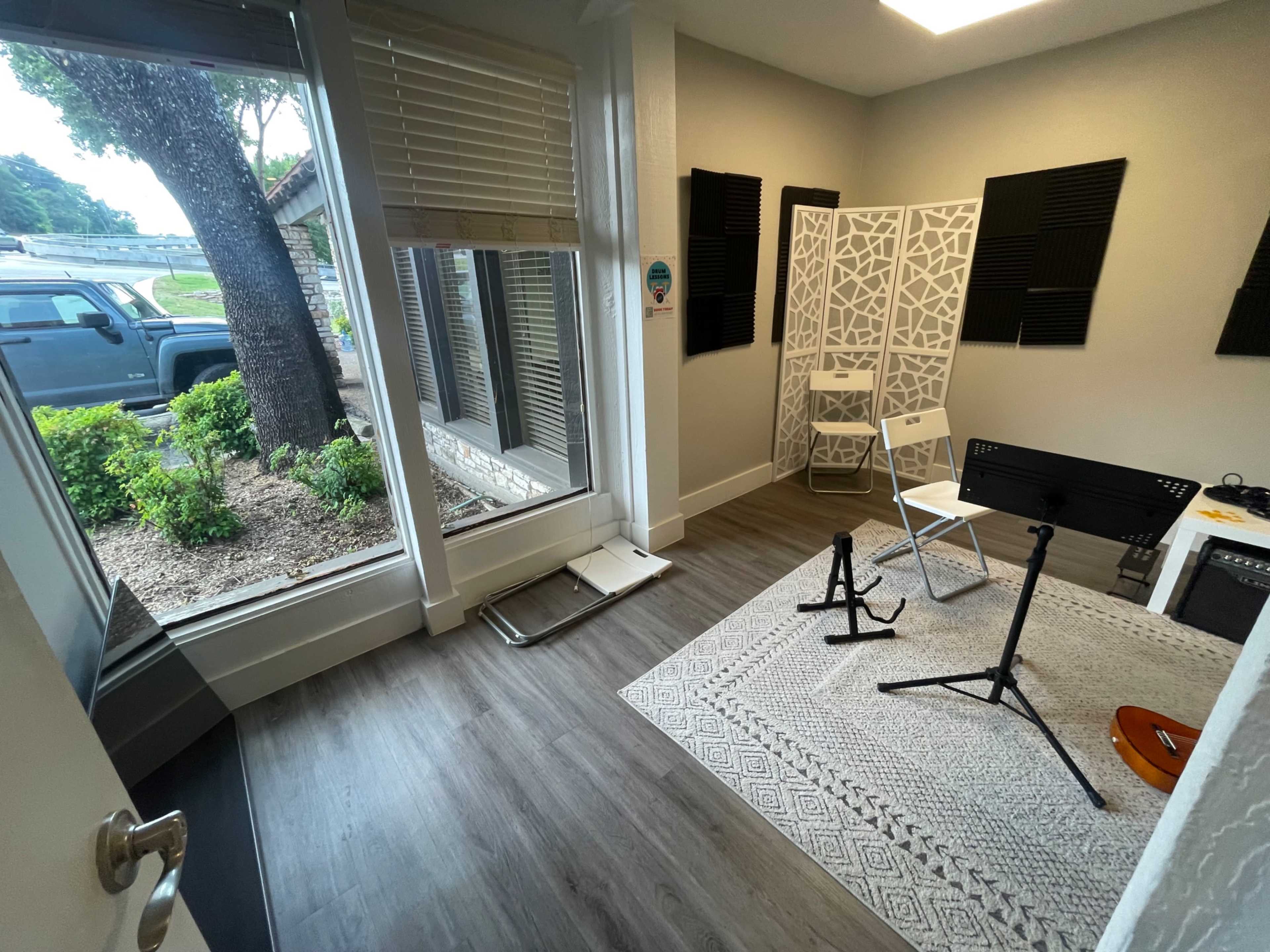 A small room with a patterned rug, chairs, and music stands, featuring soundproofing panels on the walls and a view of a tree and parking area through a large window.