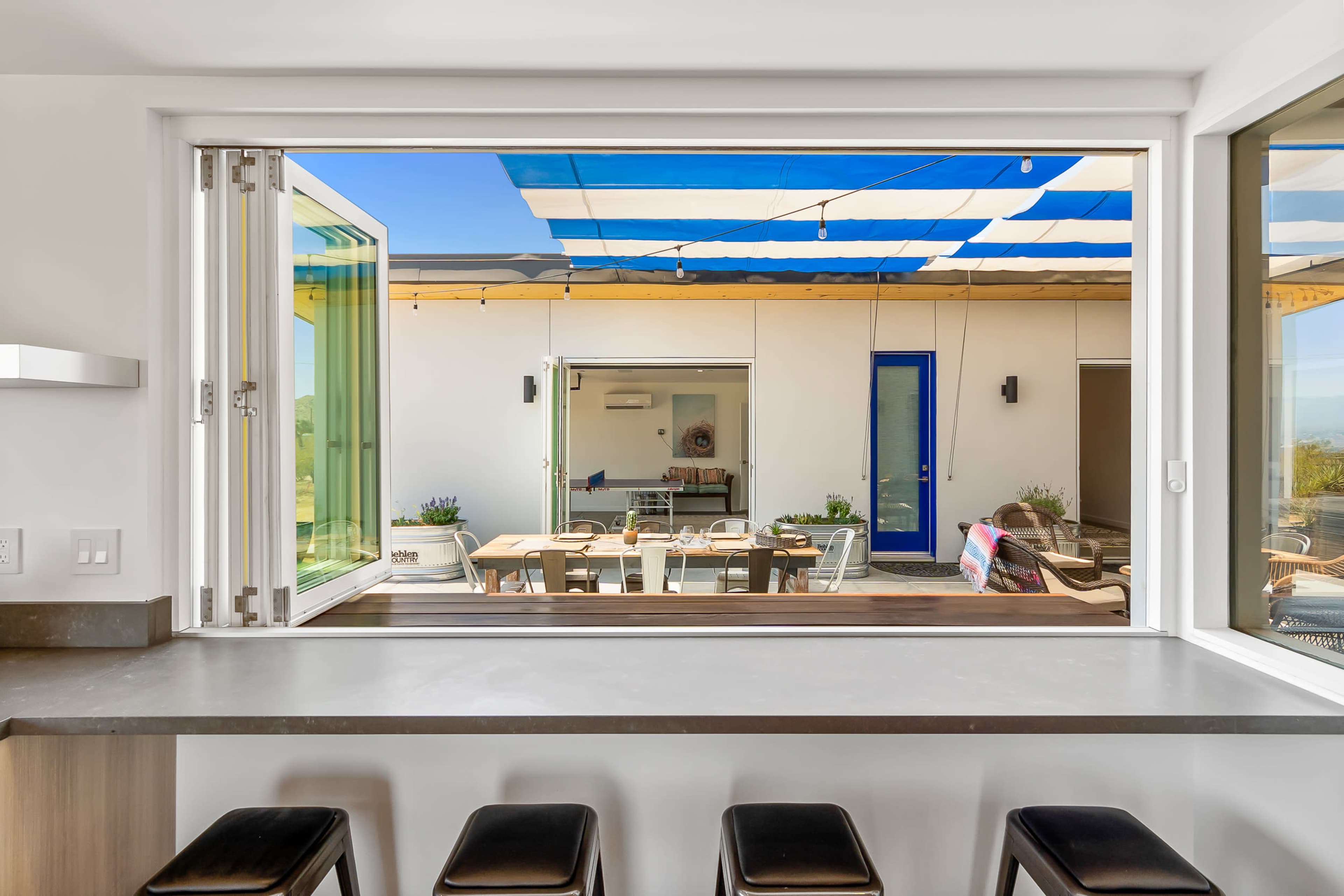 A modern kitchen features an open window that looks out onto a patio with a dining table and blue-striped shade.