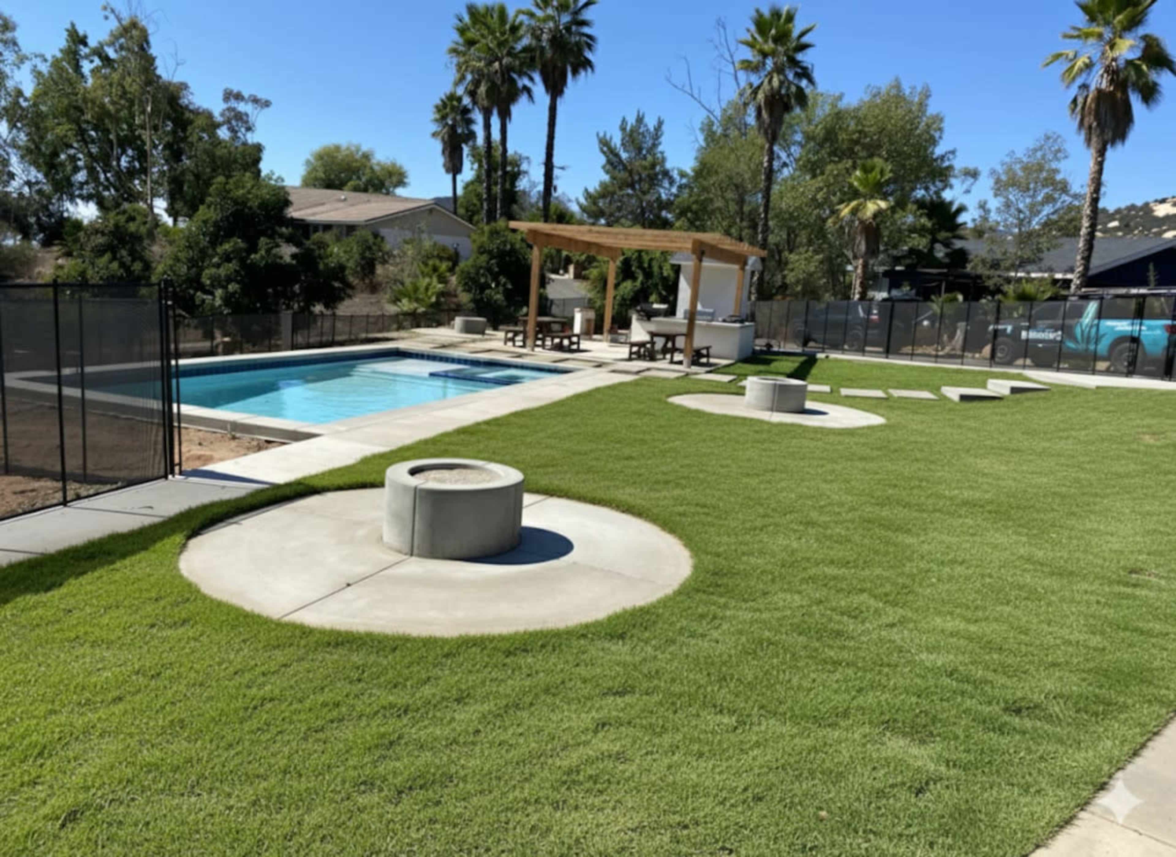 The image shows a backyard with a swimming pool, surrounded by palm trees, a green lawn, and a seating area under a wooden pergola.