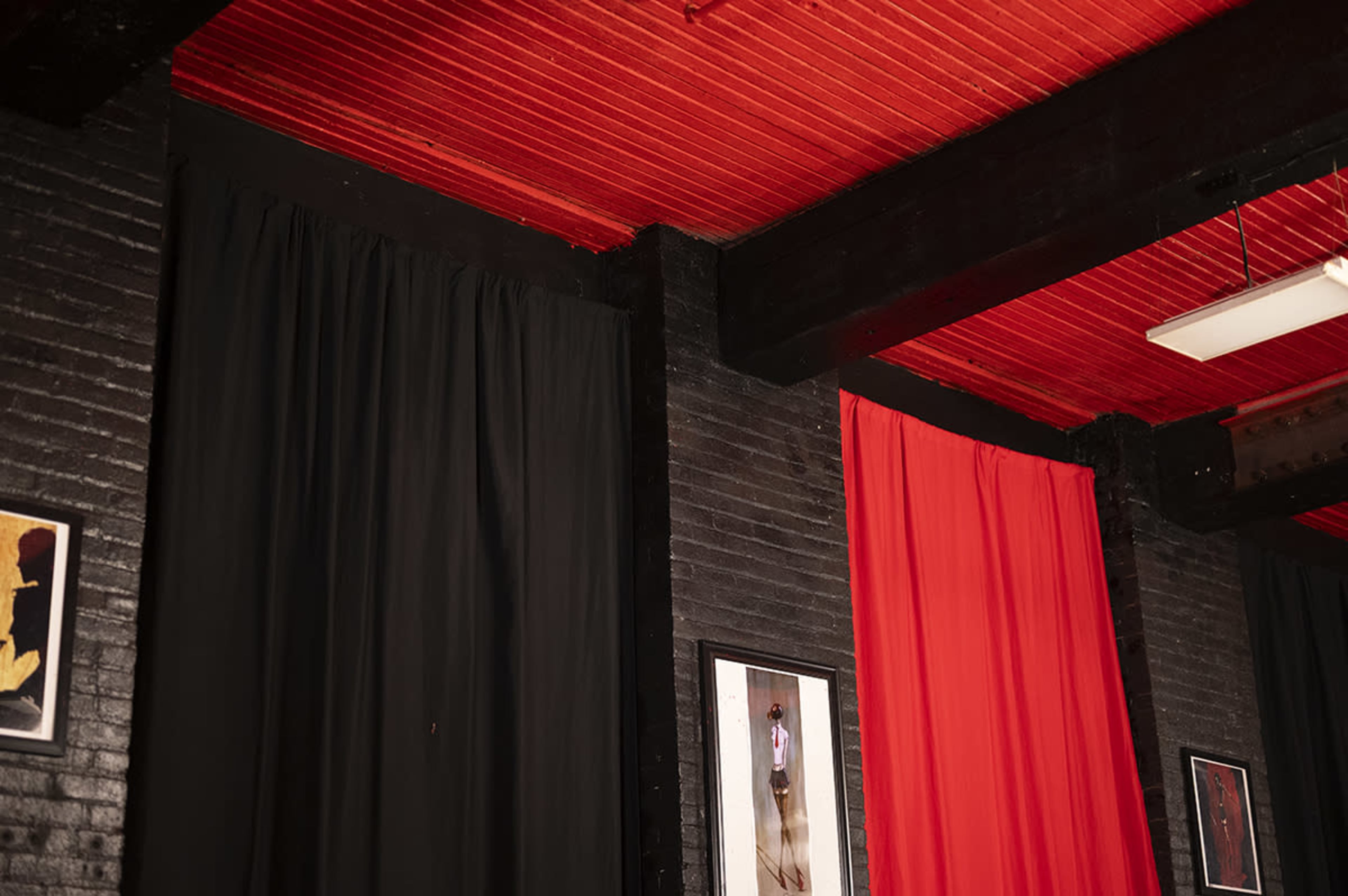 The image features a wall with black and red curtains, along with framed artworks hanging on the dark-colored brick.