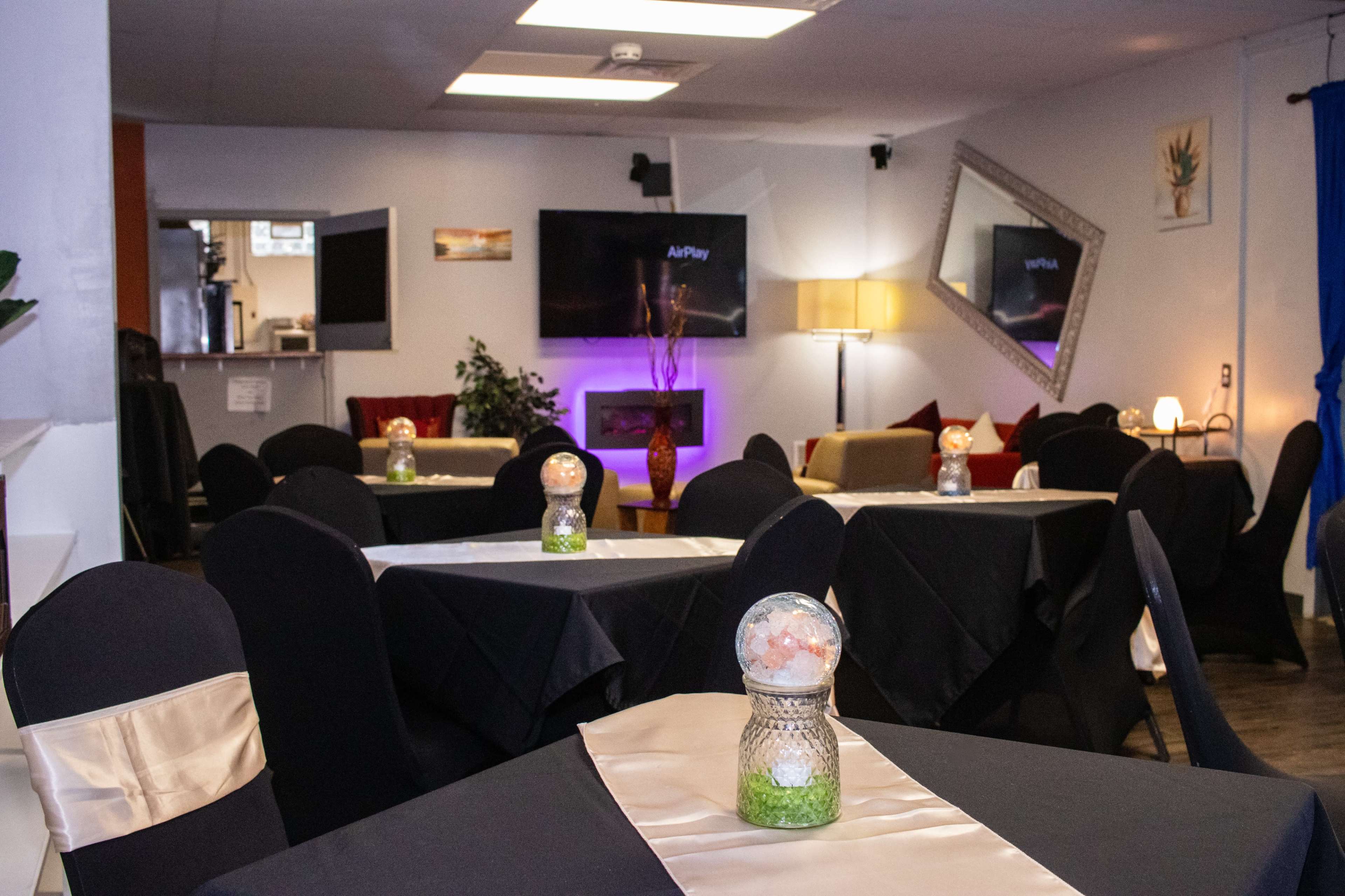 A restaurant interior with dark tables covered in black cloths and decorative centerpieces, featuring a TV, a lamp, and mirrors on the walls.