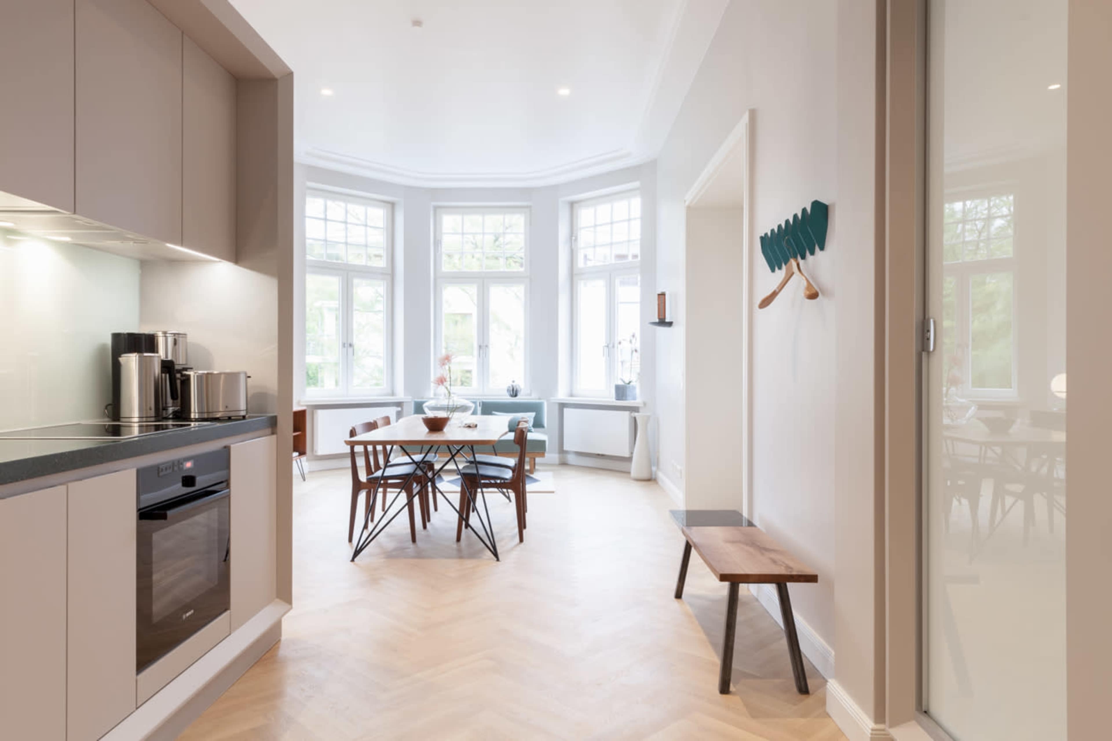 A bright, modern kitchen opens into a dining area with large windows and a wooden table surrounded by chairs.
