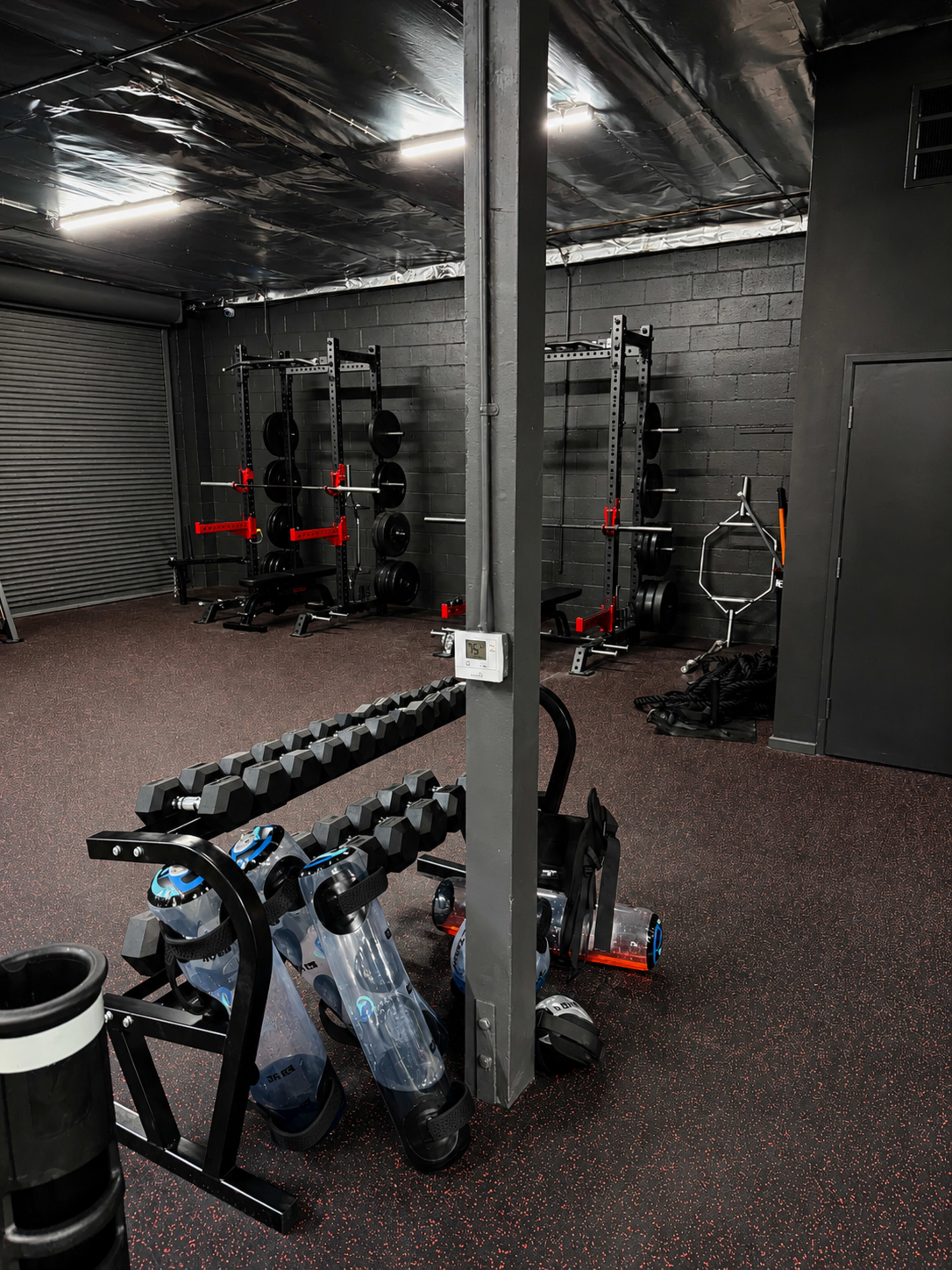 The image shows a gym interior with weightlifting equipment, including a rack of dumbbells and a barbell setup, against a black wall and flooring made of rubber material.
