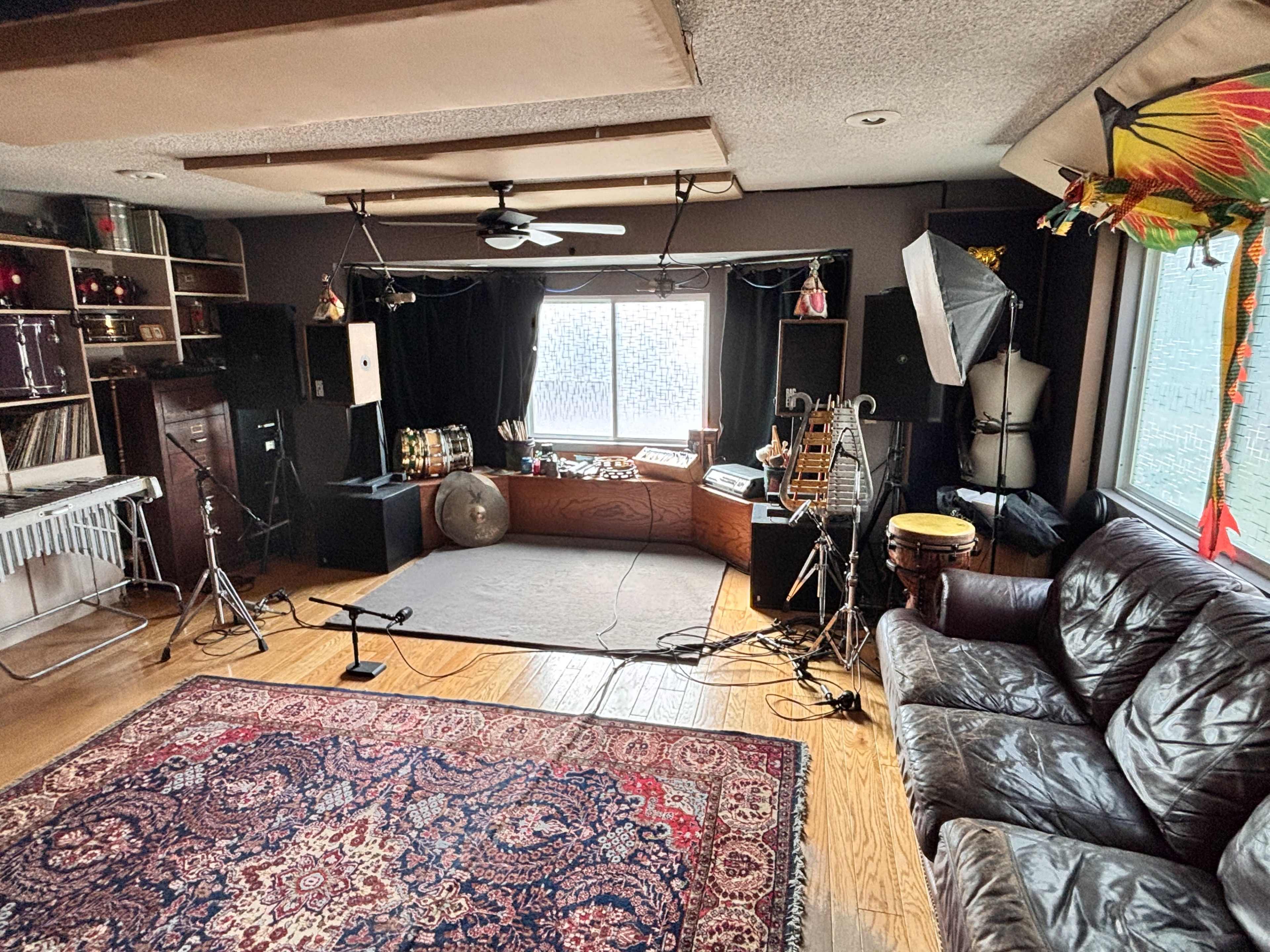 The image shows a music studio featuring a variety of instruments, microphones, and recording equipment arranged around a central performance area, with a large window providing natural light.