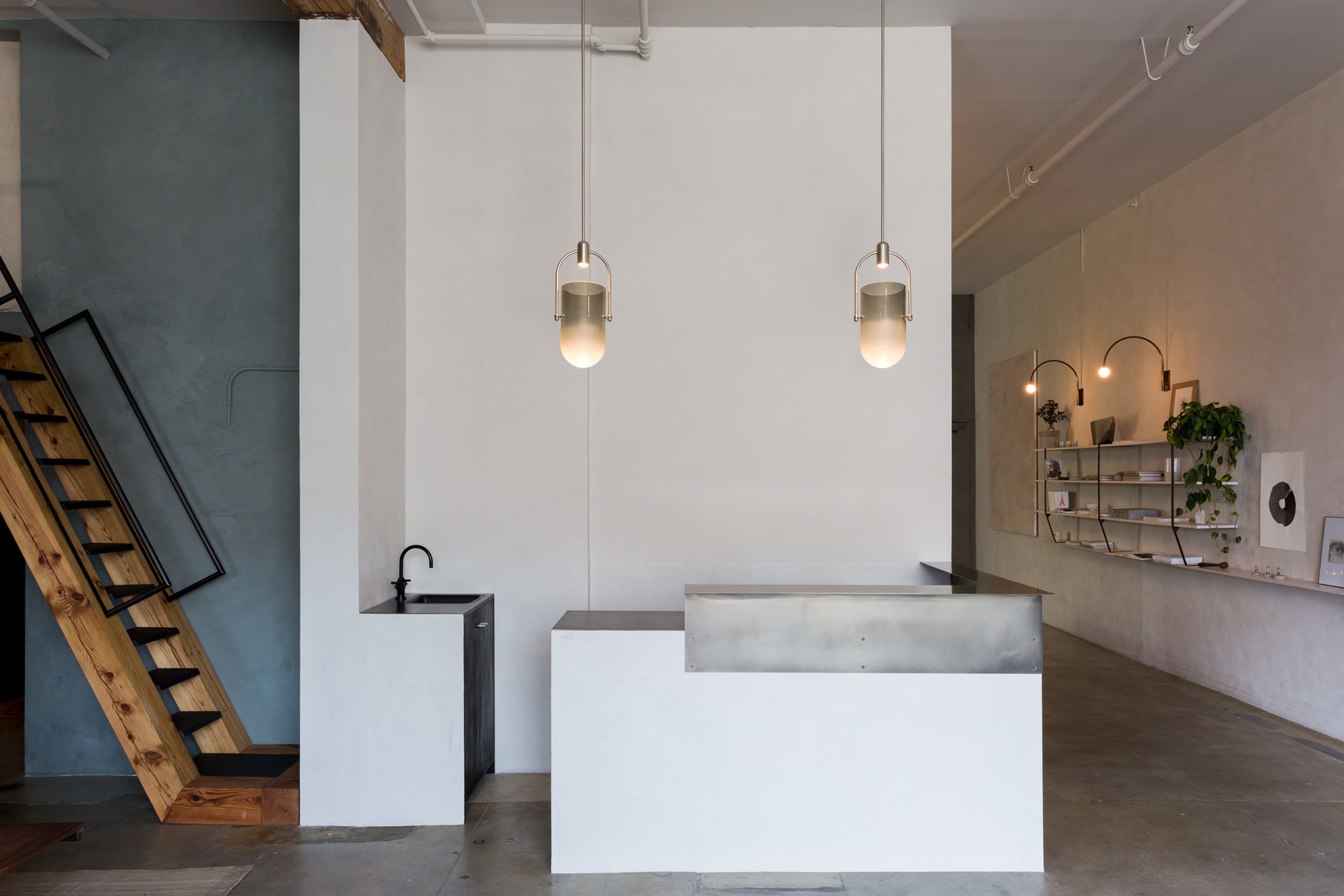 The image shows a minimalist interior with a small kitchen area featuring a metallic countertop and a sink, adjacent to a wooden staircase and shelves with plants on a light-colored wall.