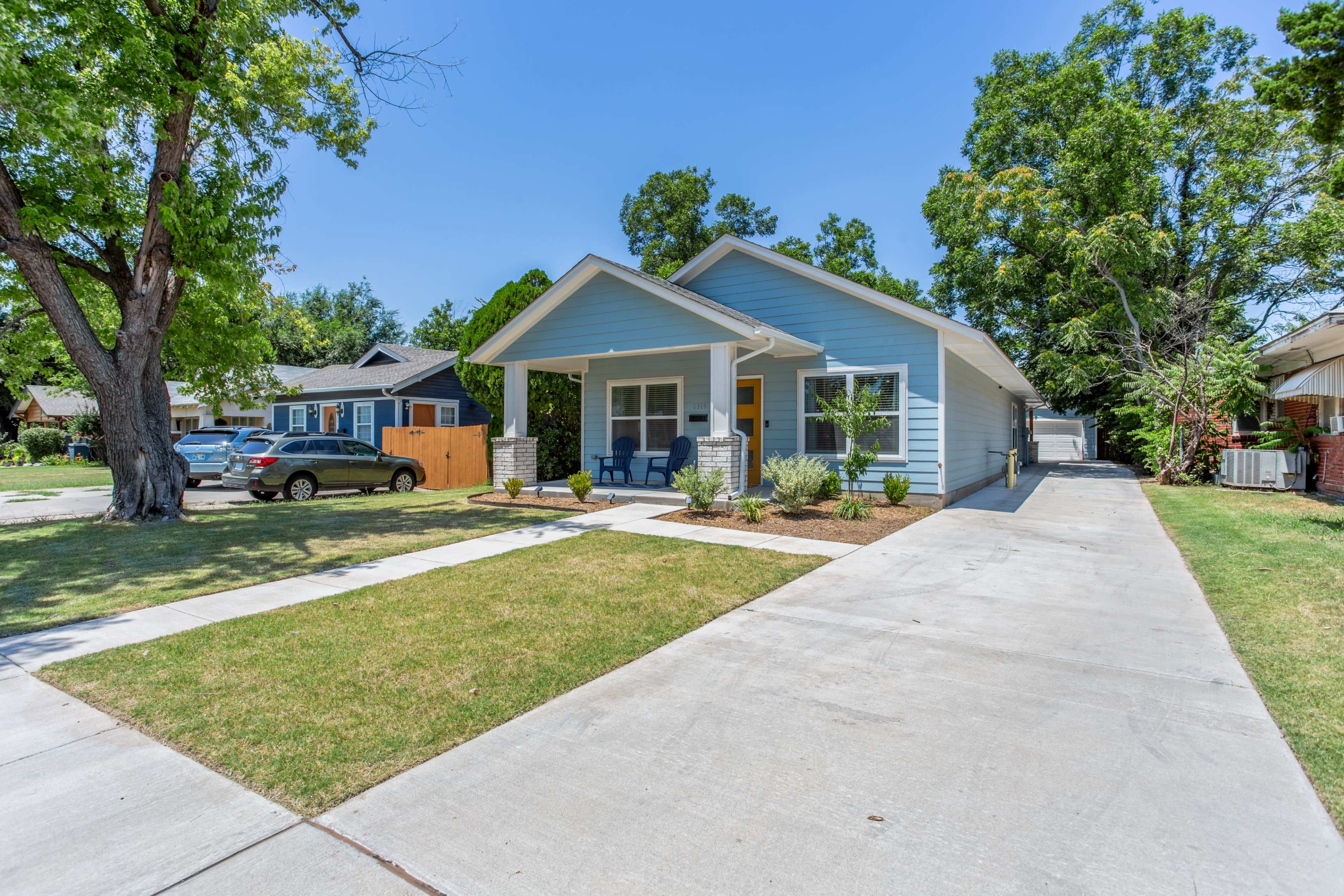 A light blue single-story house with a front porch is situated on a landscaped lawn, flanked by trees on a sunny day.