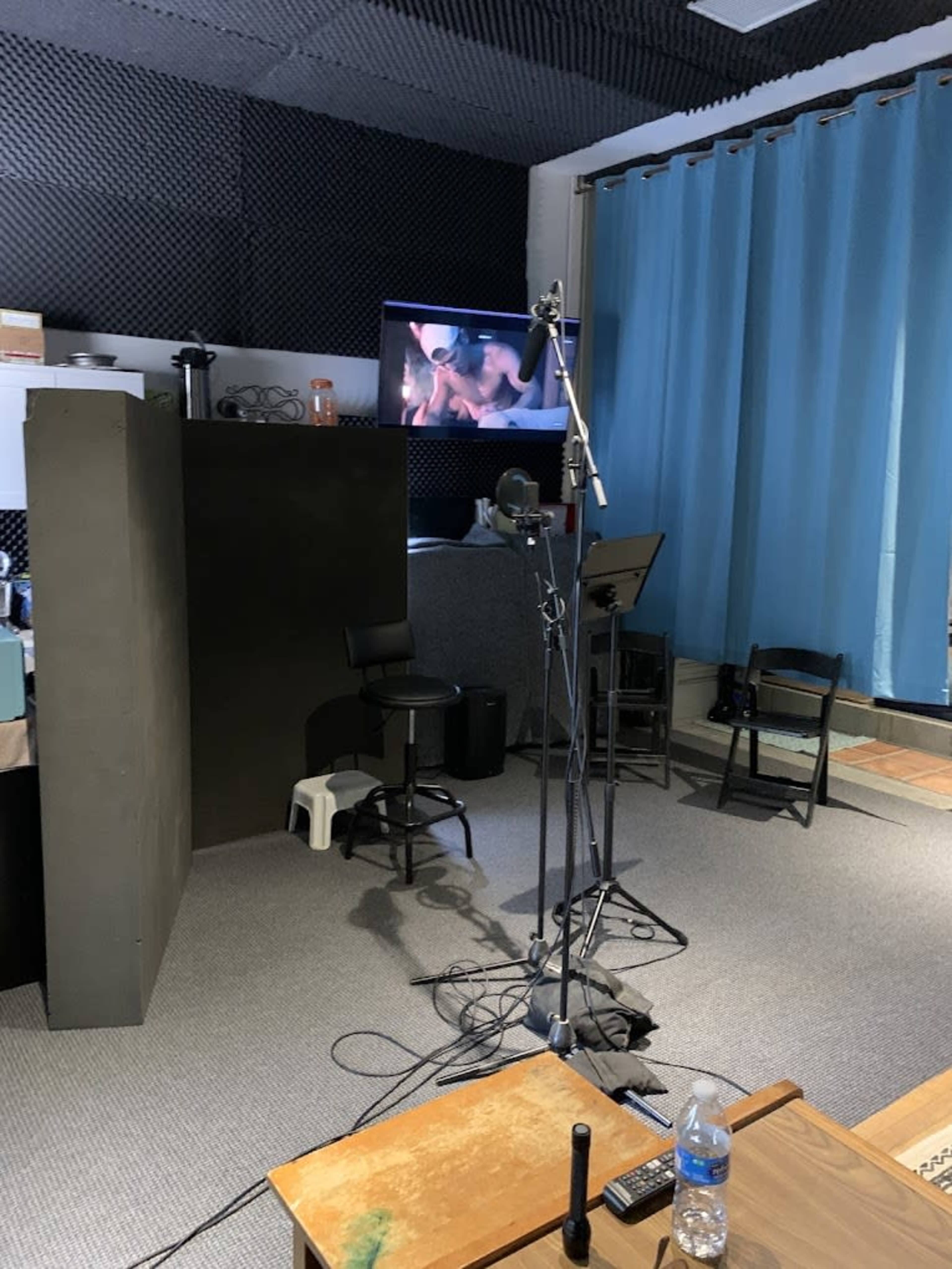 The image depicts a recording studio setup featuring a microphone on a stand, a chair, and a television screen displaying a video, with blue curtains and soundproofing materials in the background.
