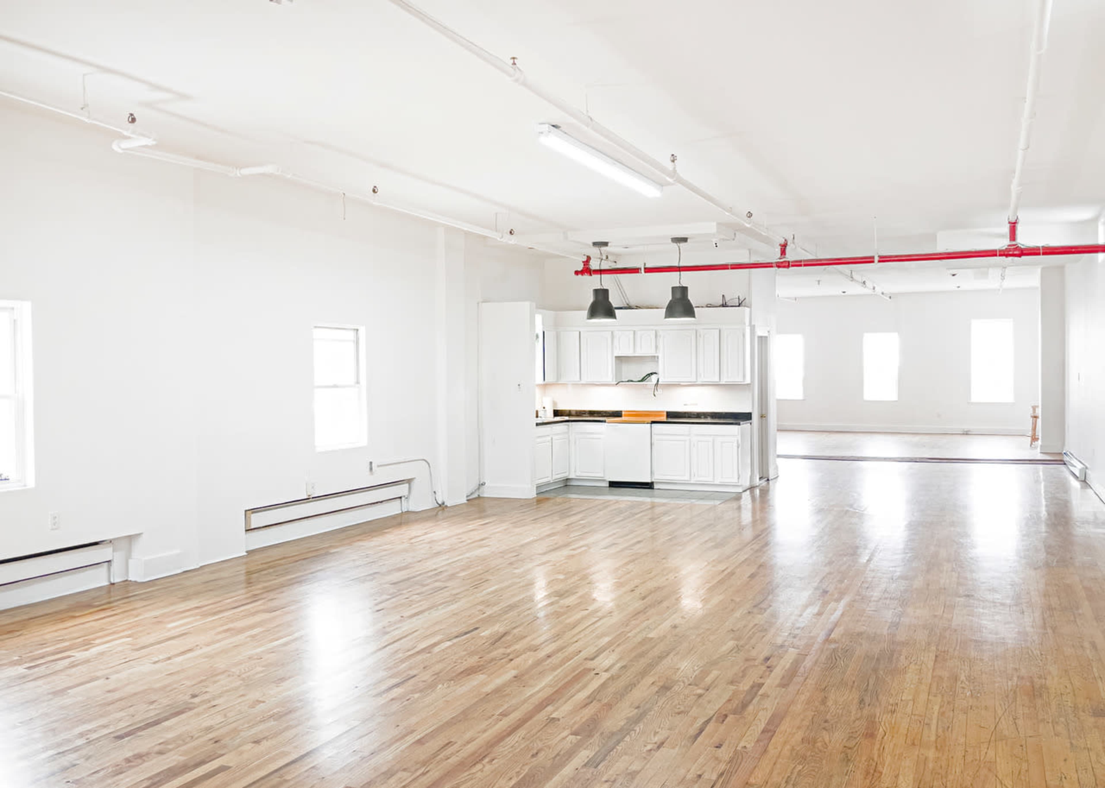 A spacious, empty room with hardwood floors, white walls, and a kitchen area featuring white cabinets and overhead lights.