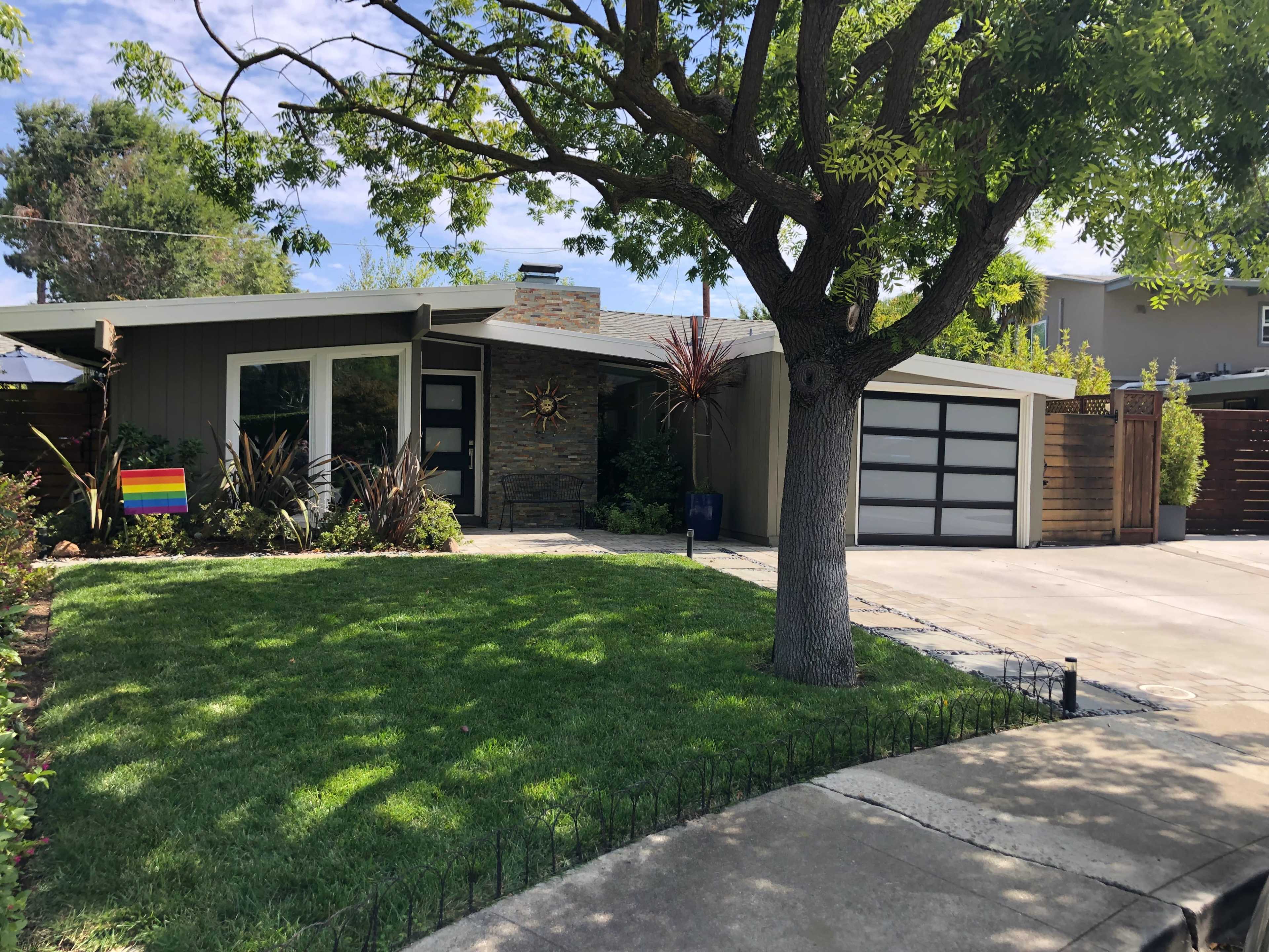 A modern house with a stone accent wall and a colorful pride flag in the yard is surrounded by well-maintained landscaping and a driveway leading to a two-car garage.