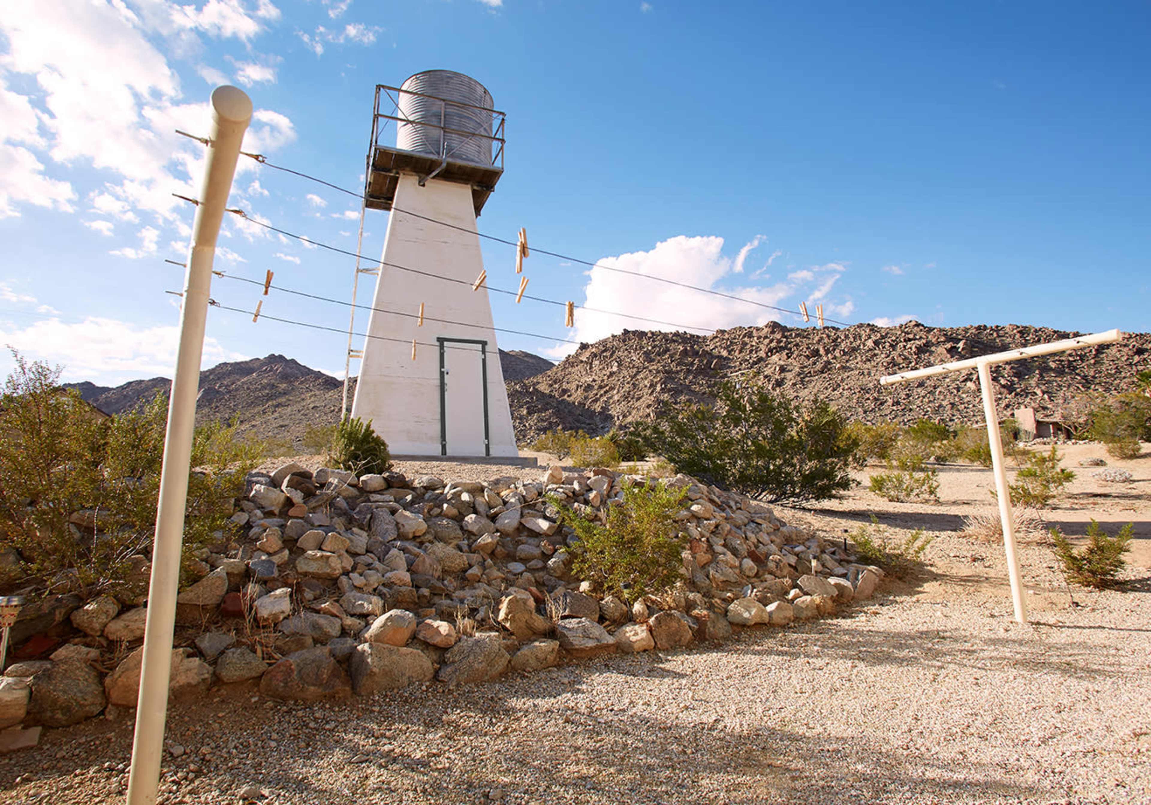 A tall water tower stands on a rocky mound with a clothesline in the foreground against a backdrop of hills under a blue sky.