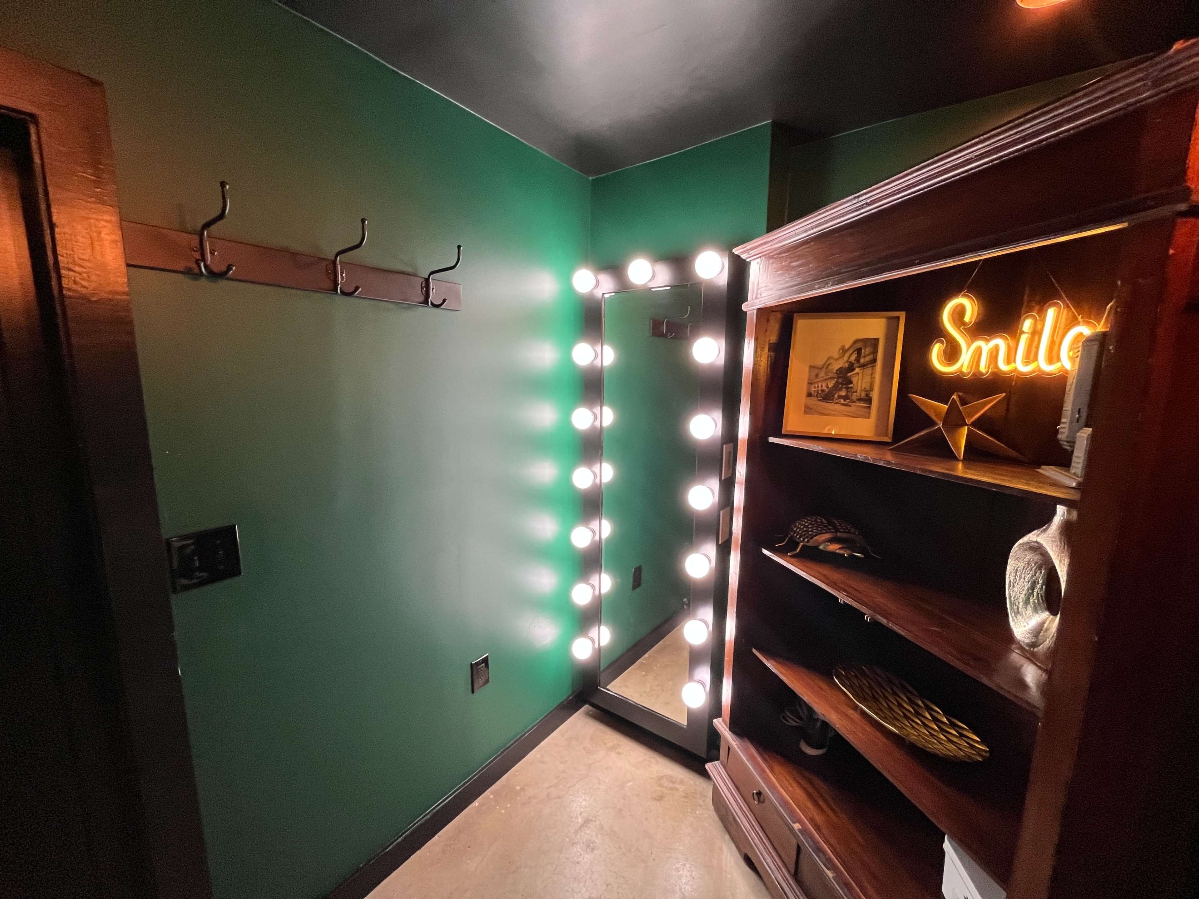 A small room with green walls, featuring a large illuminated mirror, a wooden shelf, and coat hooks on one side.