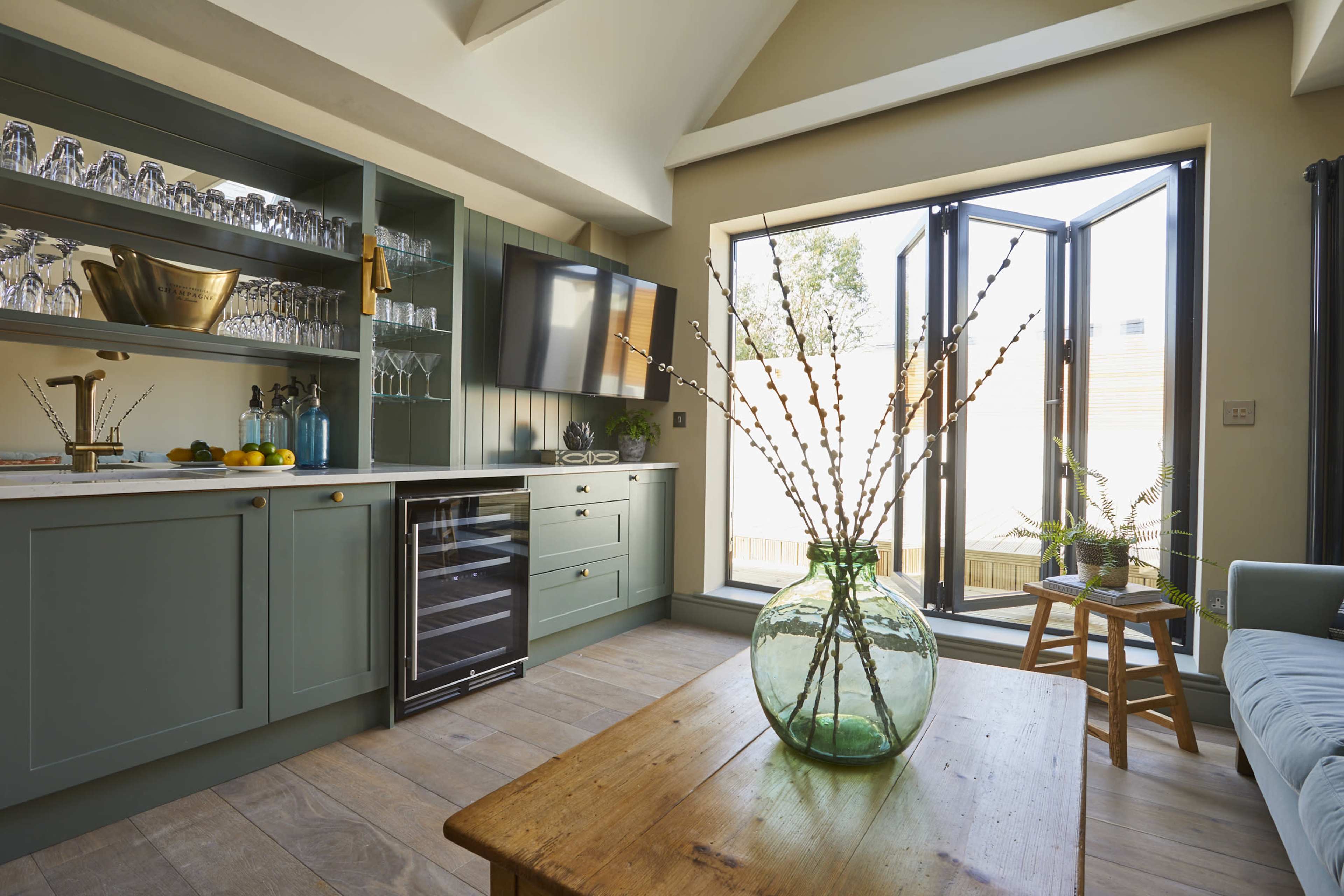 A modern kitchen features a green cabinetry setup with a wine cooler, a glassware display, and a large window that opens to an outdoor area.